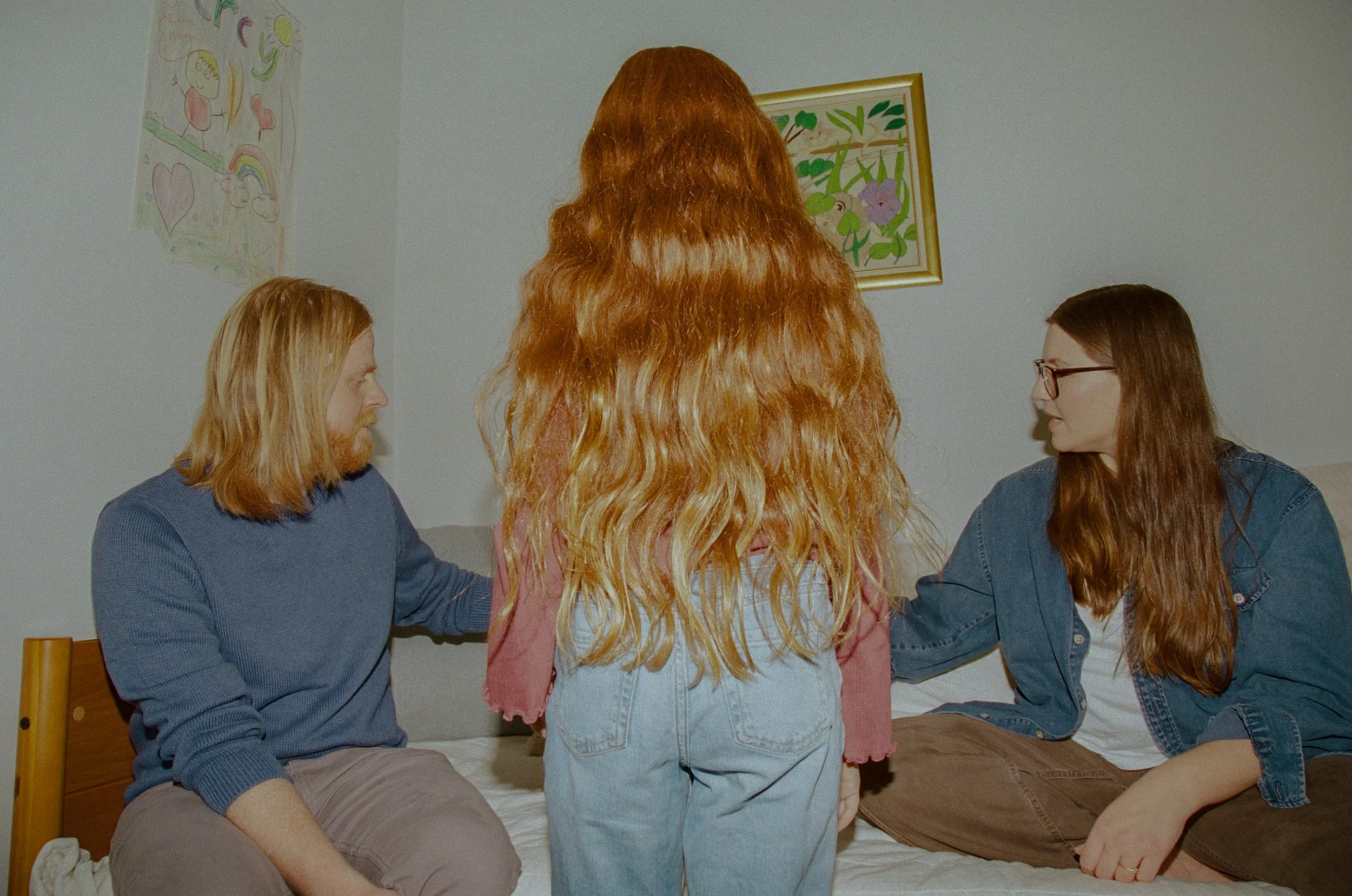 Two people sitting on a bed, facing each other, with another person standing between them. The person on the left has long red hair and is wearing a blue sweater, while the person on the right has brown hair, glasses, and is wearing a denim jacket. T