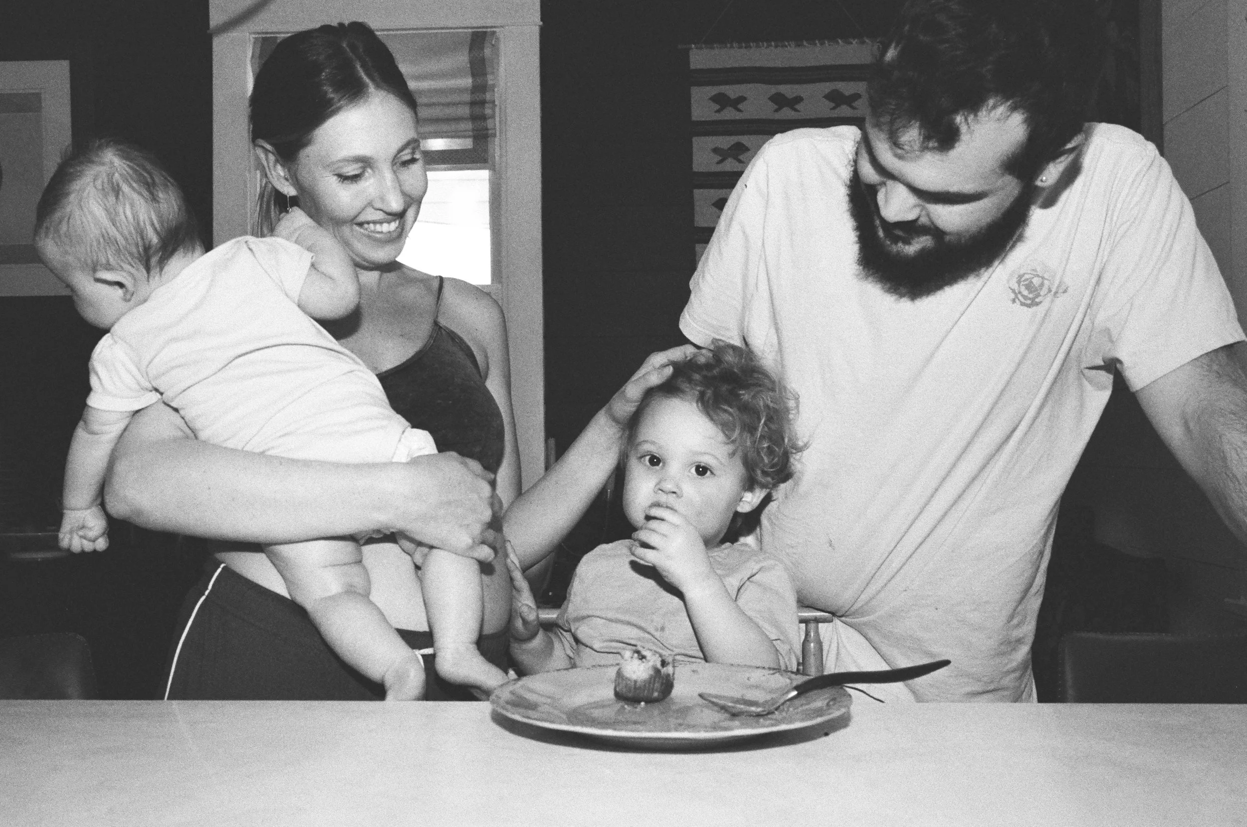 A woman holding a baby, a young boy sitting at a table with a cupcake, and a man leaning over them, all smiling or looking at the camera in a kitchen during their postpartum newborn photoshoot in Richmond, Virginia on 35mm film
