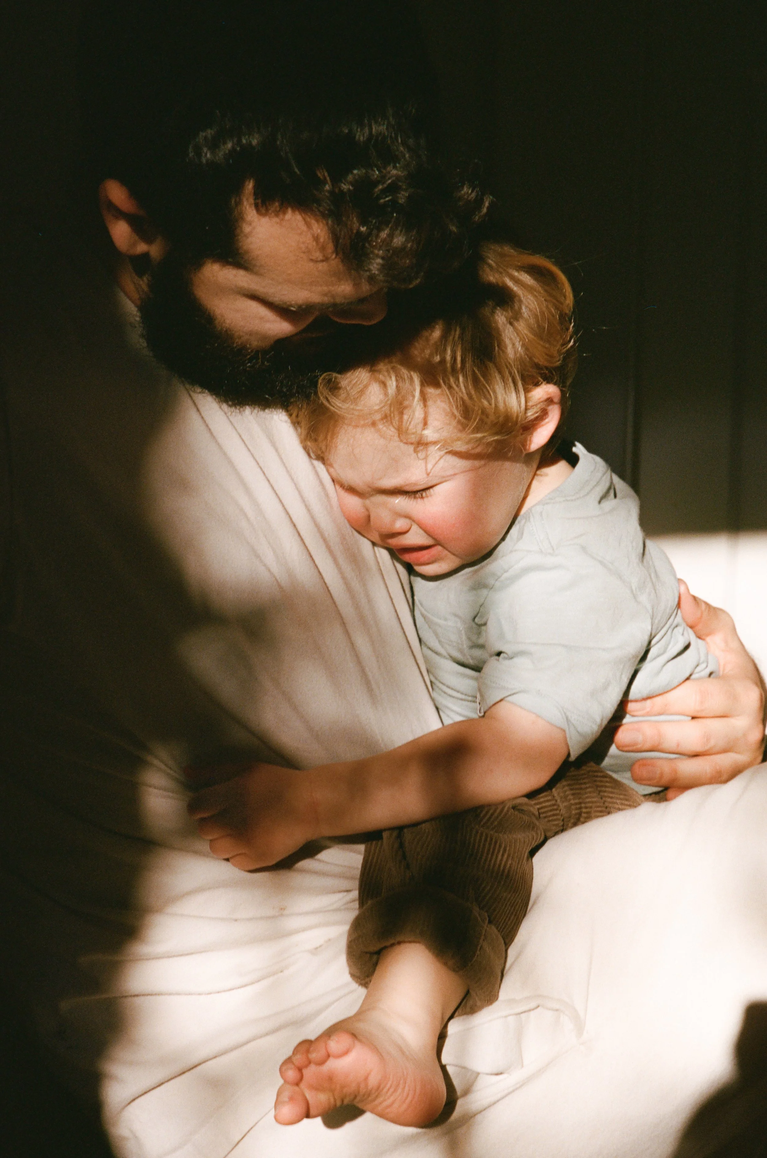 A man with dark hair and beard holding a young boy, who appears upset or crying, in his arms. The boy has light brown hair and is wearing a light gray shirt and brown pants. The scene is intimate and emotional. 35mm film natural light. 