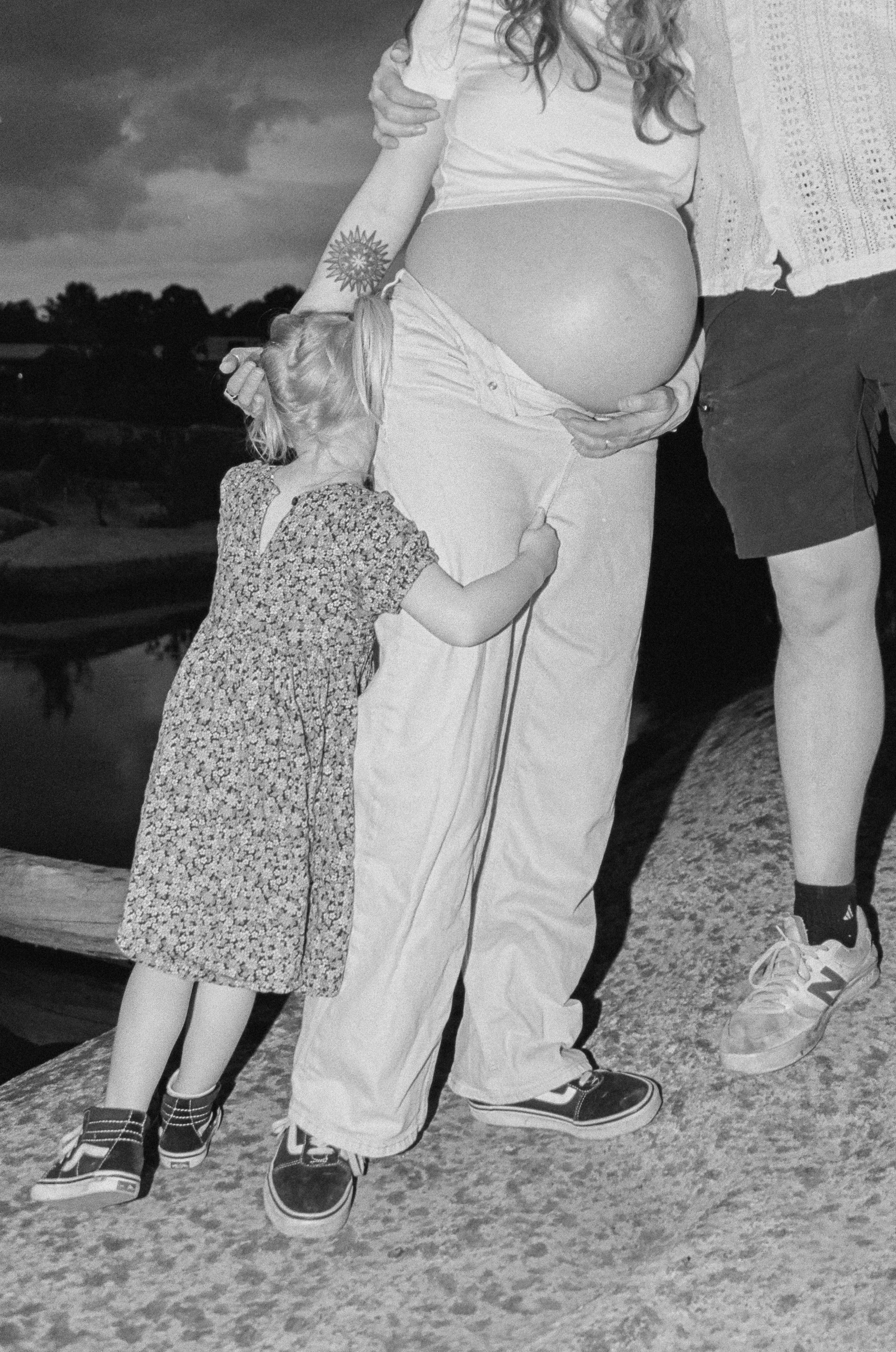A black and white photo of a pregnant woman, standing outdoors, gently holding her belly with both hands. A young girl is hugging her, with her head resting on the woman's belly. The woman is wearing a casual white T-shirt and light-colored pants, and the girl is wearing a patterned dress and sneakers. There are people in the background and a body of water, indicating an outdoor or park setting.