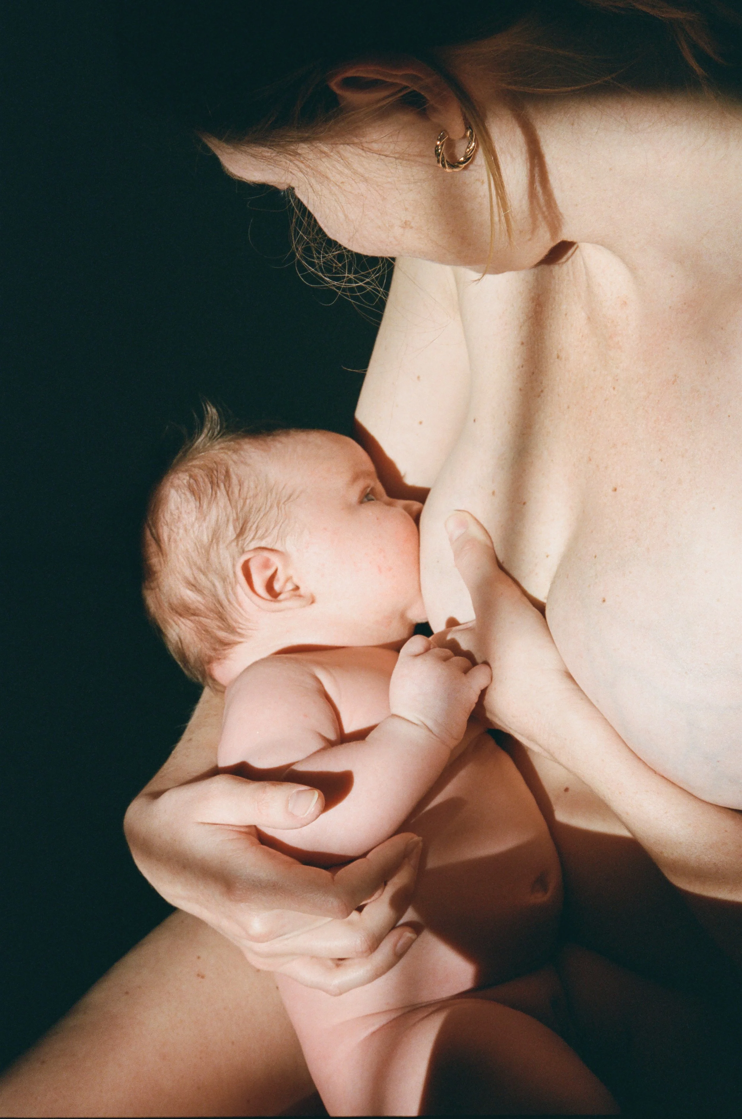 A mother breastfeeding her baby against a moody dark background, showing the baby latched onto her breast photographed on 35mm film in natural sunlight. 