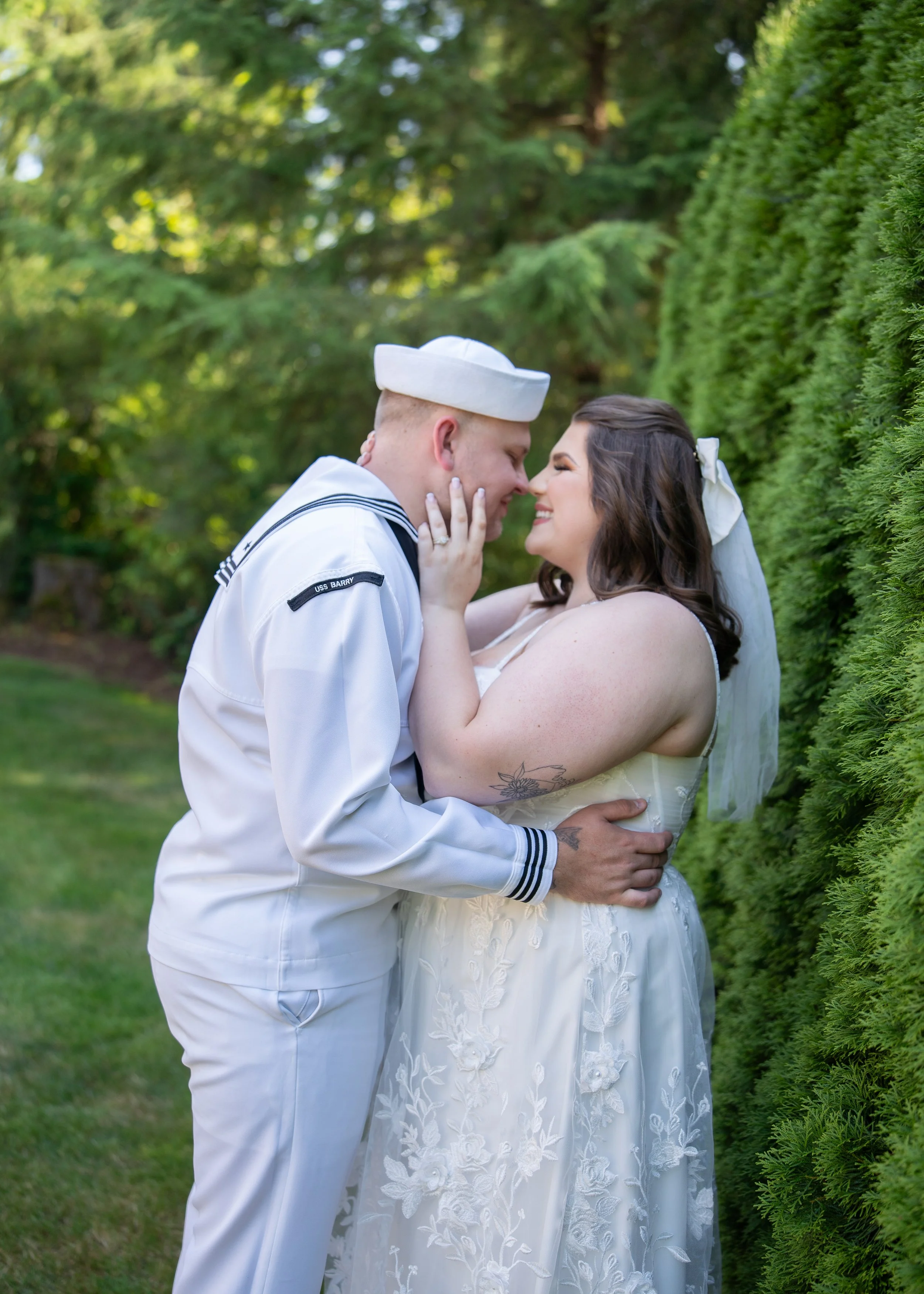 A bride and groom, dressed in wedding attire, embrace and smile as they lean close to each other outdoors surrounded by greenery.