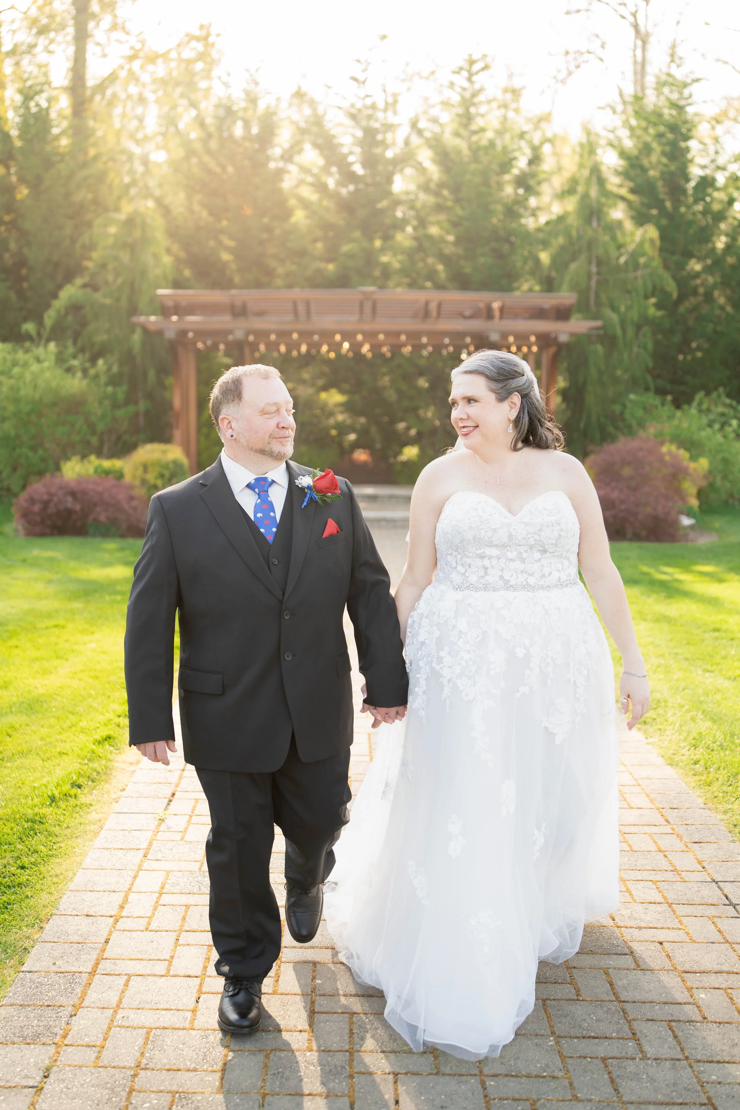 A bride and groom walking hand in hand on a brick pathway during sunset at an outdoor wedding venue.