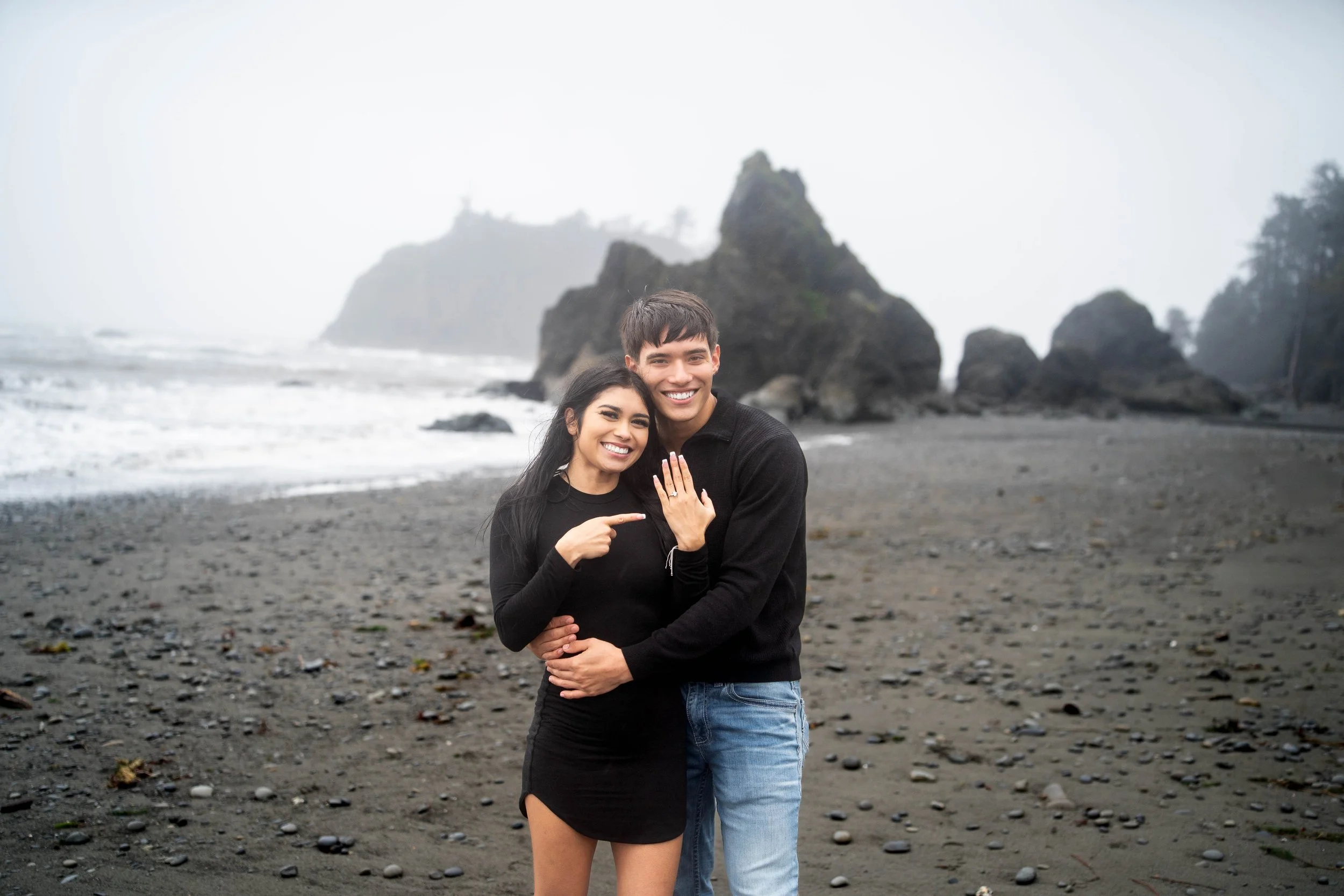 A happy couple stands on a misty, rocky beach with large rocks in the background. The woman points to her engagement ring while the man embraces her, both smiling.