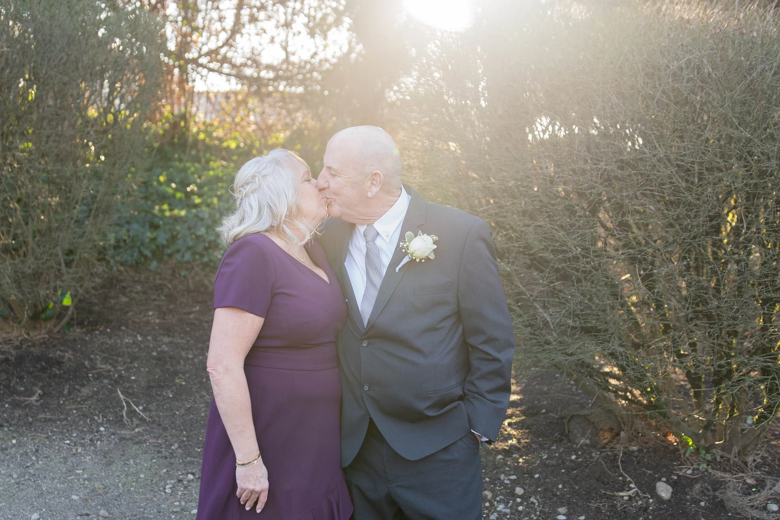 An elderly couple sharing a kiss outdoors during sunset, dressed in formal attire, with the woman wearing a purple dress and the man in a dark suit with a white boutonniere.