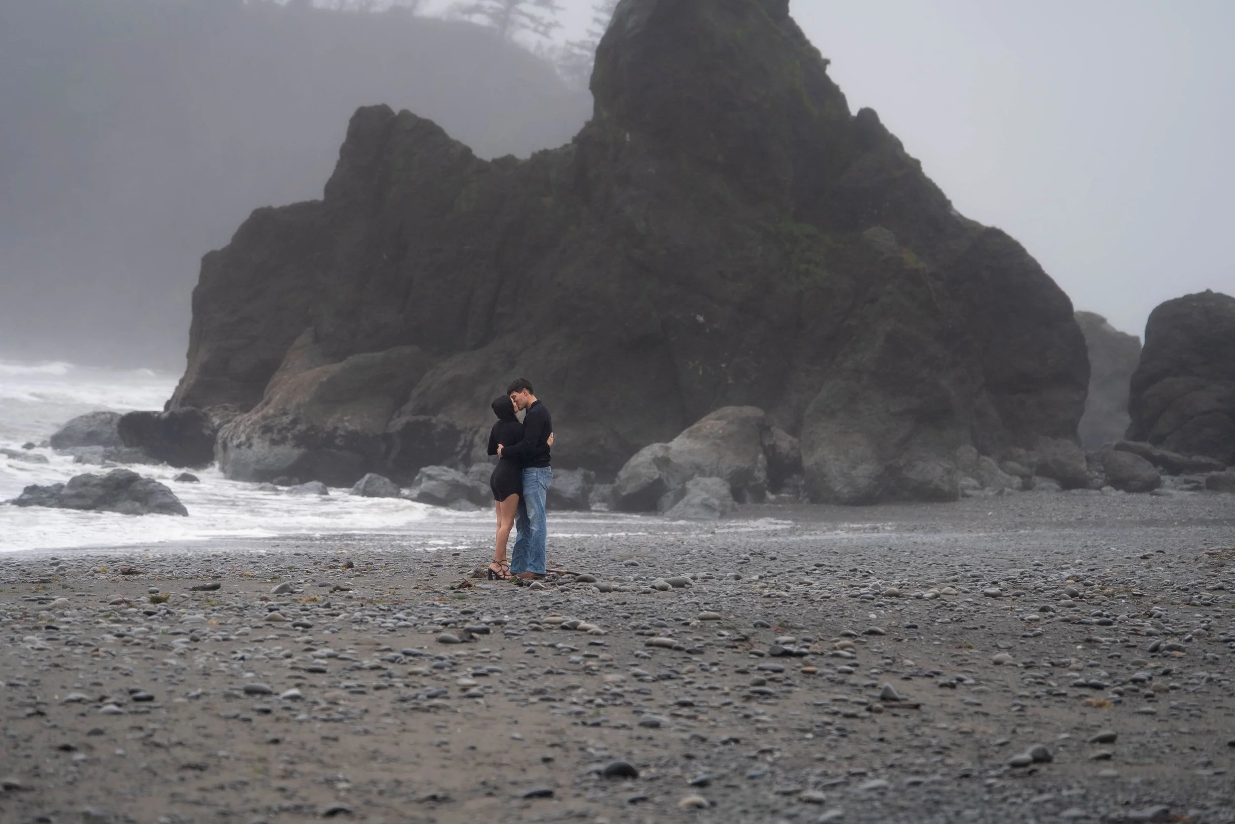 A couple embracing on a foggy rocky beach with large cliffs in the background.