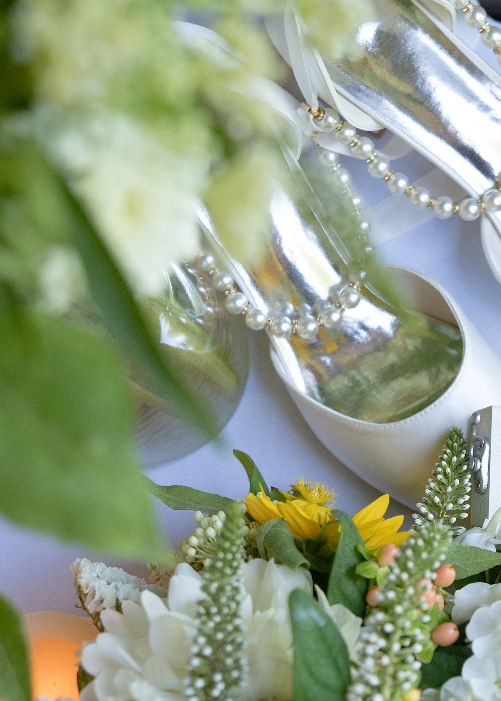 A close-up of white ballet flats with pearl and silver bead embellishments, surrounded by a floral arrangement including white, yellow, and green flowers and foliage.