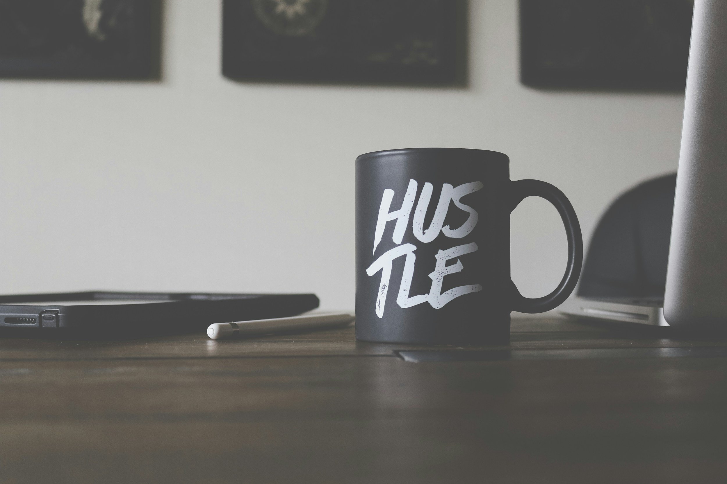 A black ceramic mug with the words 'Hustle' written in white on it placed on a wooden desk next to a closed laptop and a stylus.