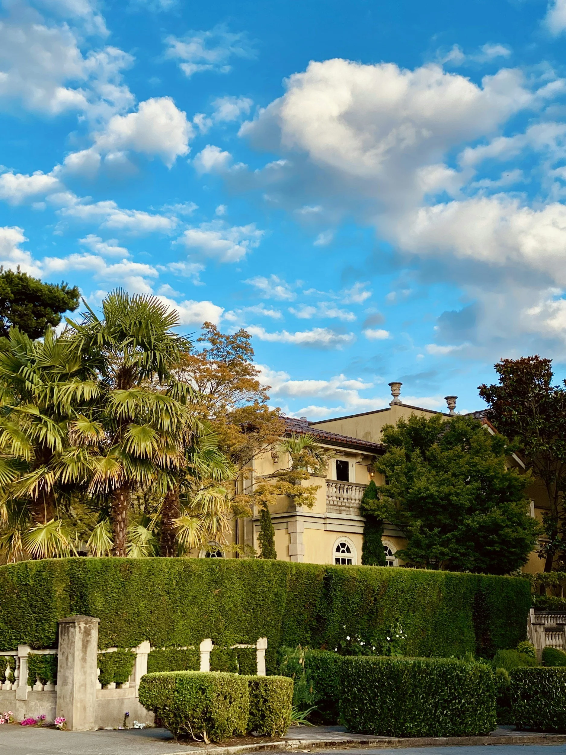 A large luxurious house with a beige exterior and brown roof, surrounded by lush green trees, manicured bushes, and palm trees, under a bright blue sky with fluffy white clouds.