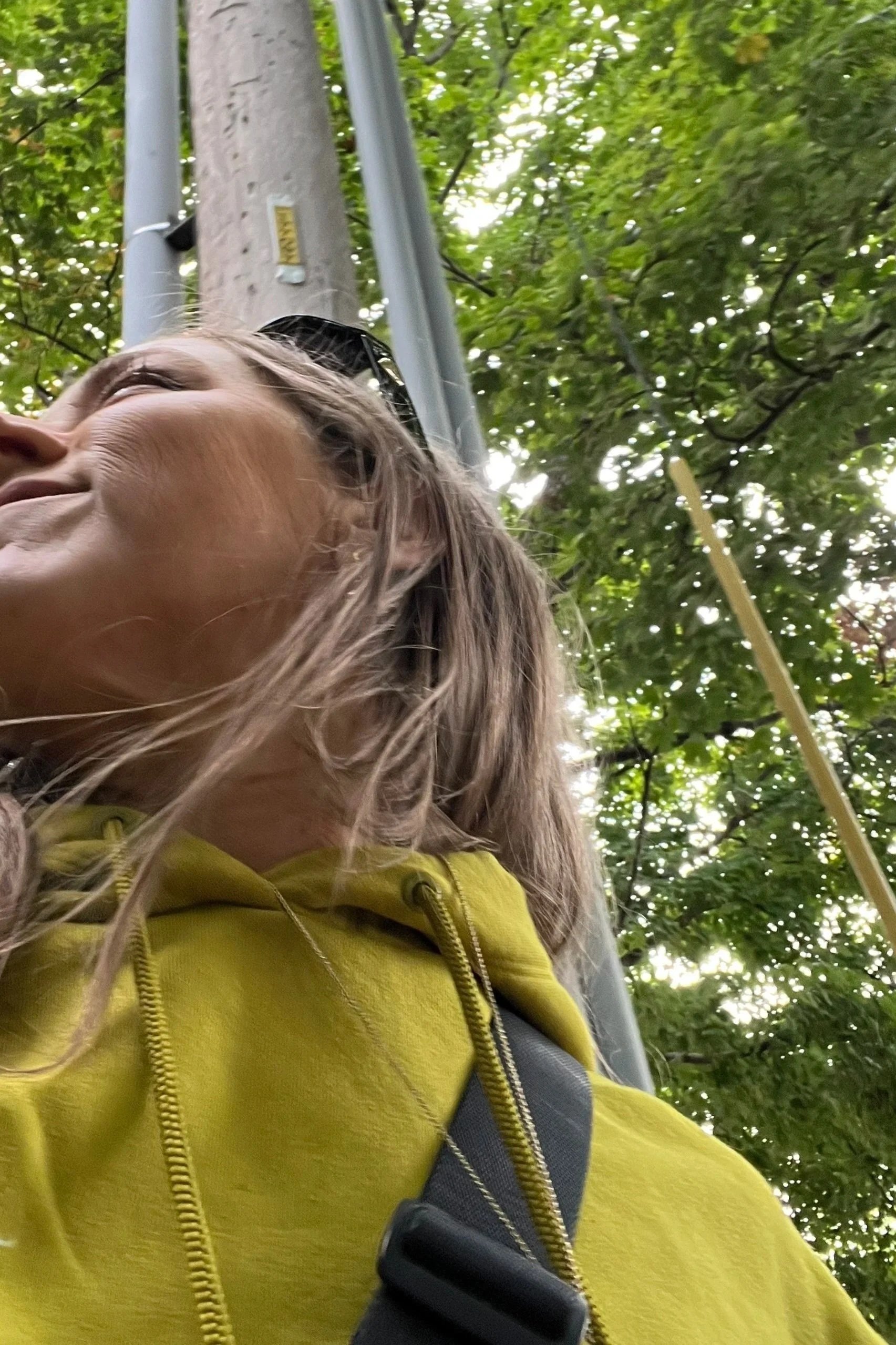 Close-up of a woman with blonde hair wearing a yellow hoodie, looking up at trees with green leaves.