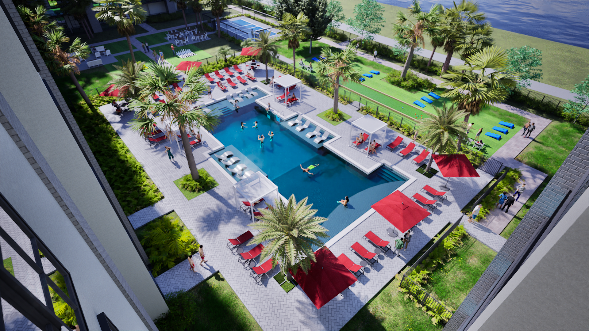 Aerial view of a luxurious apartment complex pool area with lounge chairs, umbrellas, palm trees, and people swimming and lounging near a river or lake.