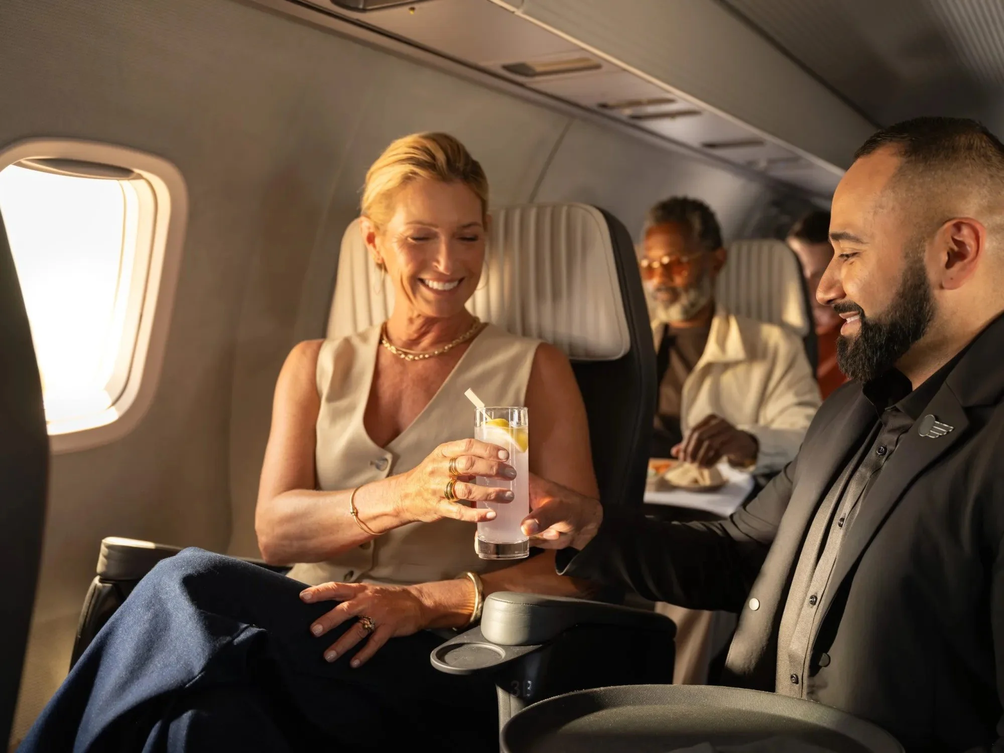 Smiling flight attendant serves a cocktail to a relaxed passenger in a spacious, upscale airplane cabin, highlighting Aero’s premium in-flight experience.