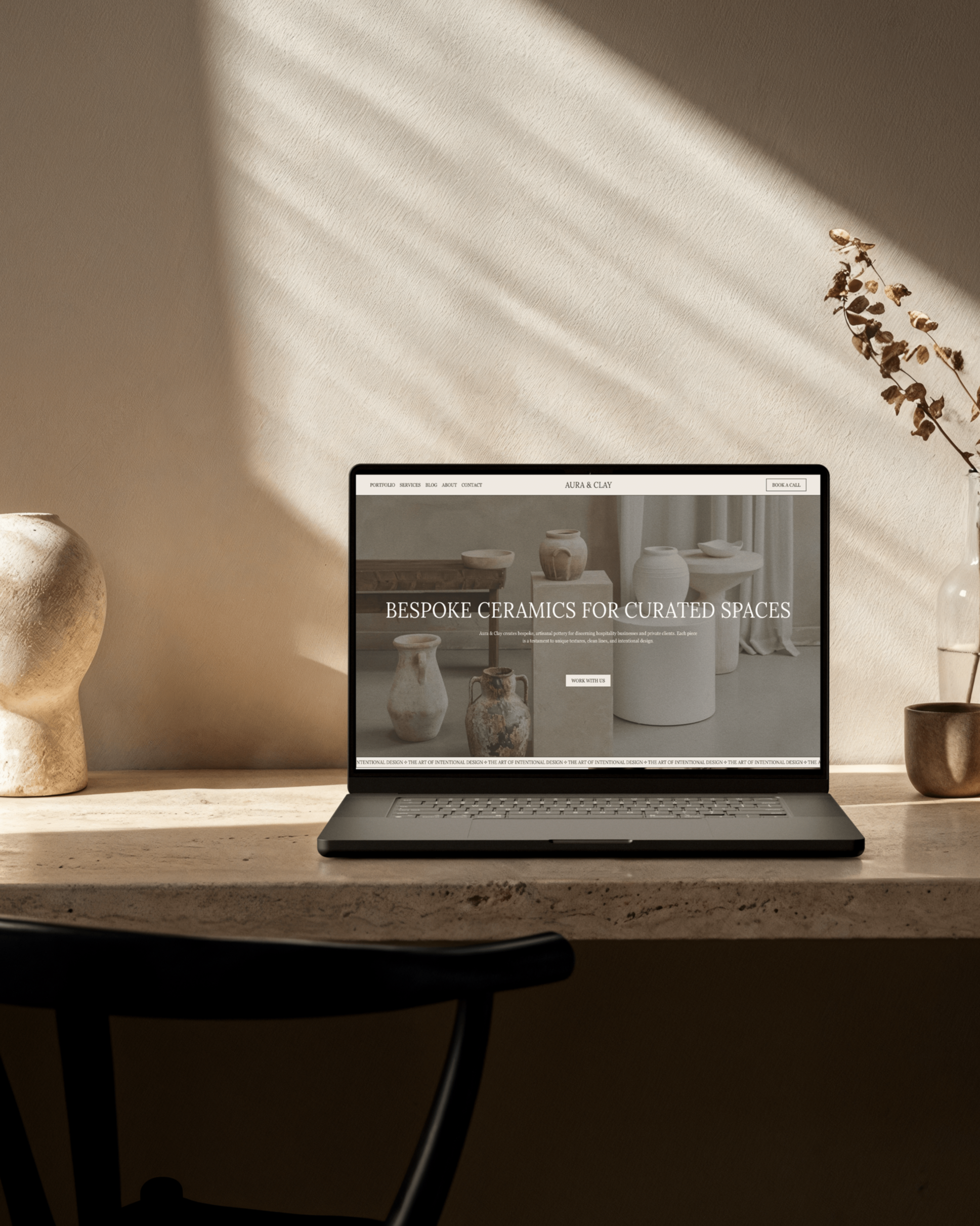 Laptop on a marble desk displaying a ceramics website with neutral-colored vases and pots in the background. Decor items and a branch in a vase are also on the desk, with sunlight casting shadows on the wall.