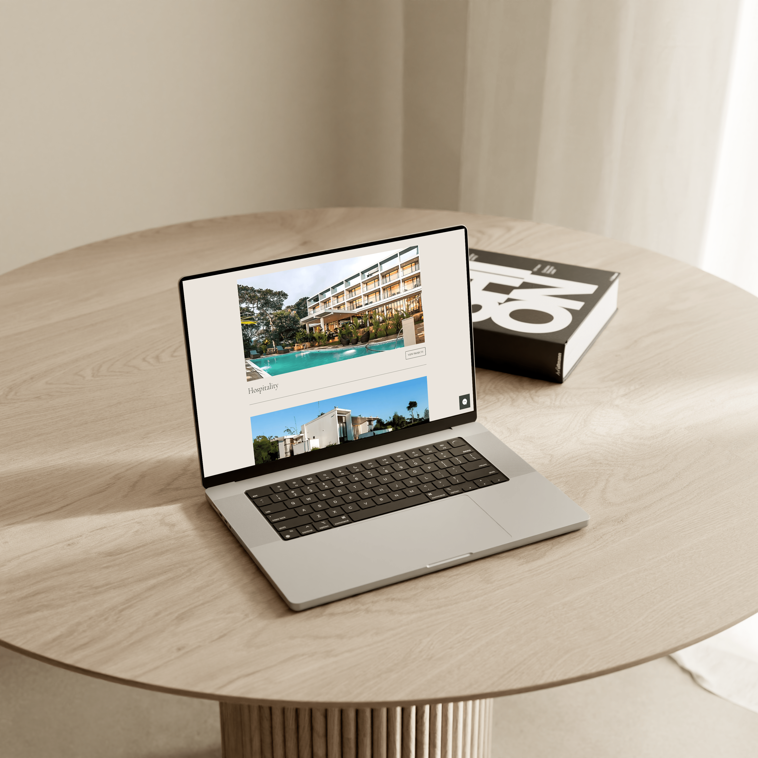 A laptop on a round wooden table displaying photos of modern hotel buildings and a swimming pool, with a thick book titled 'NOW' beside it.