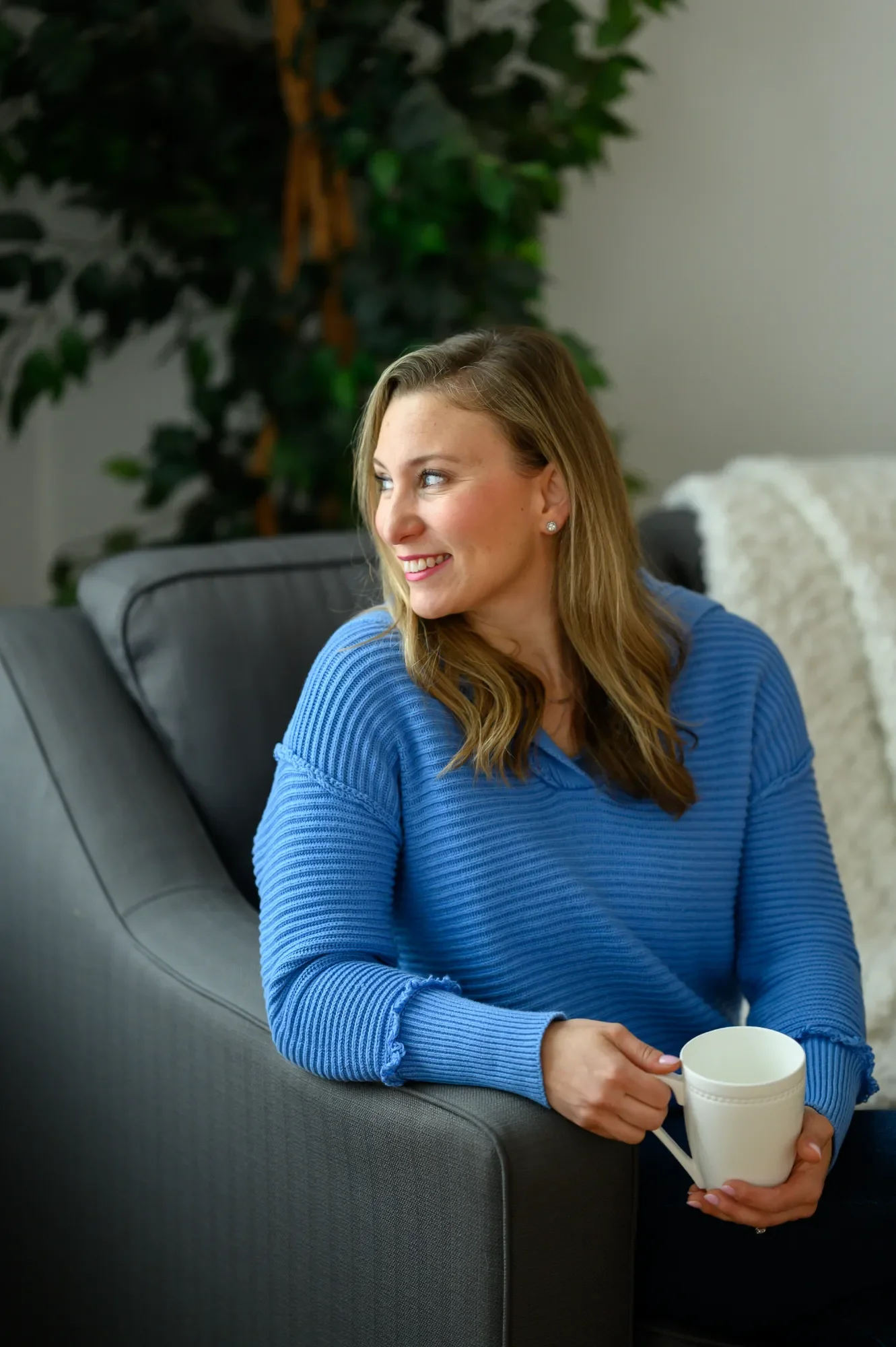 Ashley, a professional photo organizer, wearing a blue sweater, sitting on a dark gray sofa, holding a white mug, looking to her left with a smile. There is a green plant in the background.