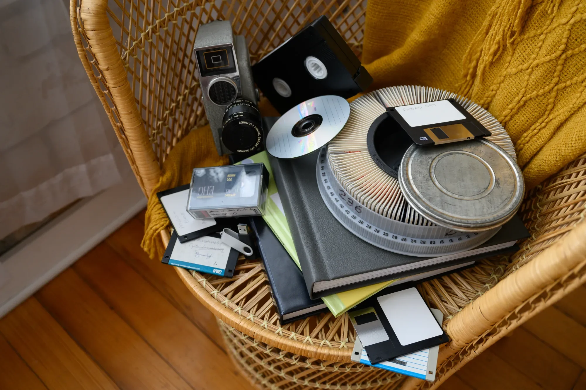 A wicker chair with a yellow blanket contains vintage film camera, floppy disks, floppy disk drive, CD, books, and film reels.