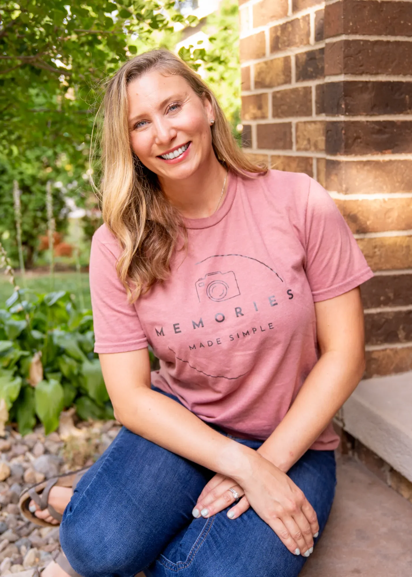 Ashley, a professional photo organizer, with long blond hair sitting outdoors near a brick wall, wearing a pink t-shirt and blue jeans.