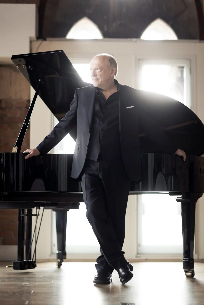 A man in a black suit stands next to a grand piano with one hand resting on the piano and the other on his hip, in a bright room with large windows.