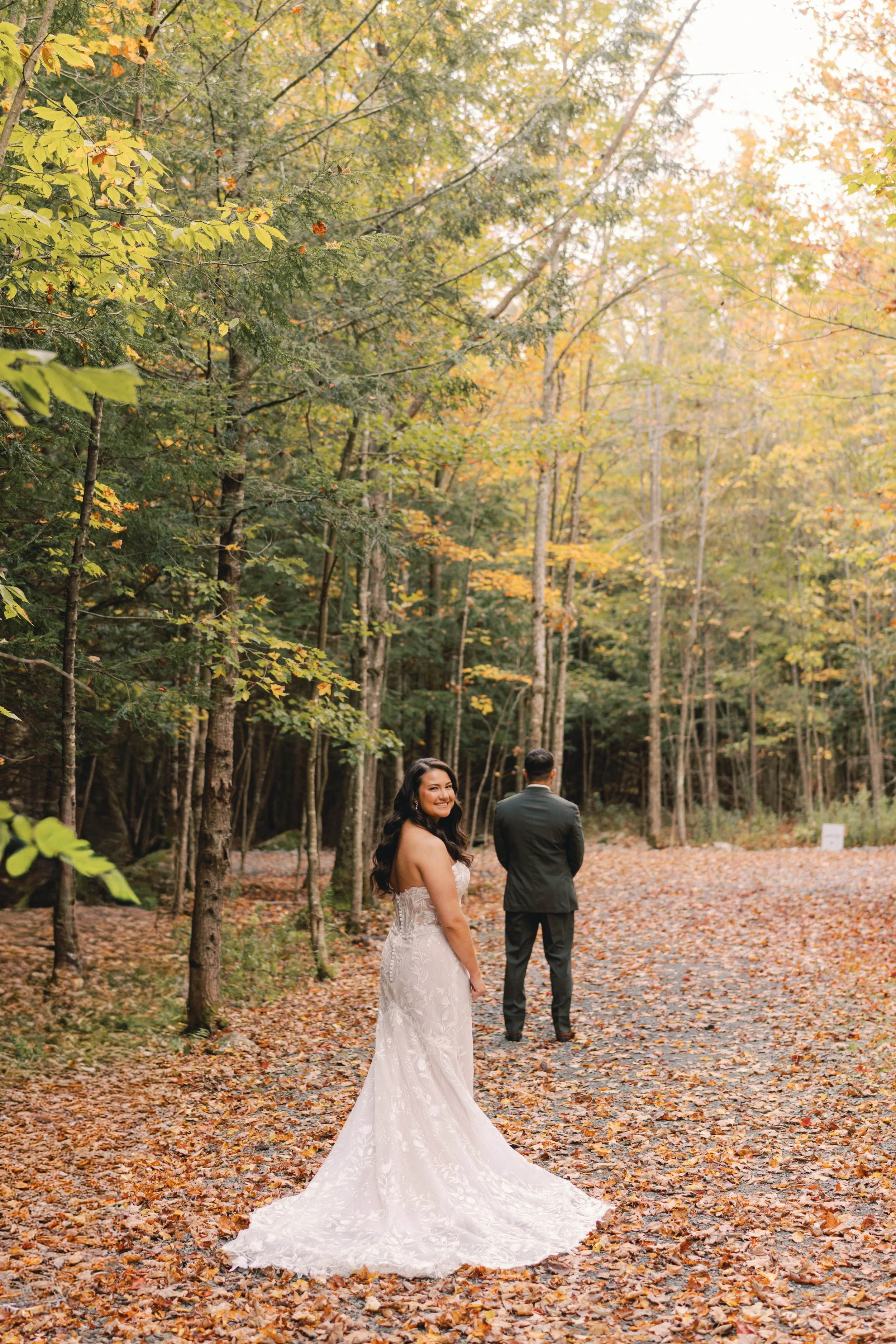 A bride in a white wedding dress with a long train standing in a wooded area with autumn leaves, smiling and looking back at the camera, with a groom in a suit facing away in the background.
