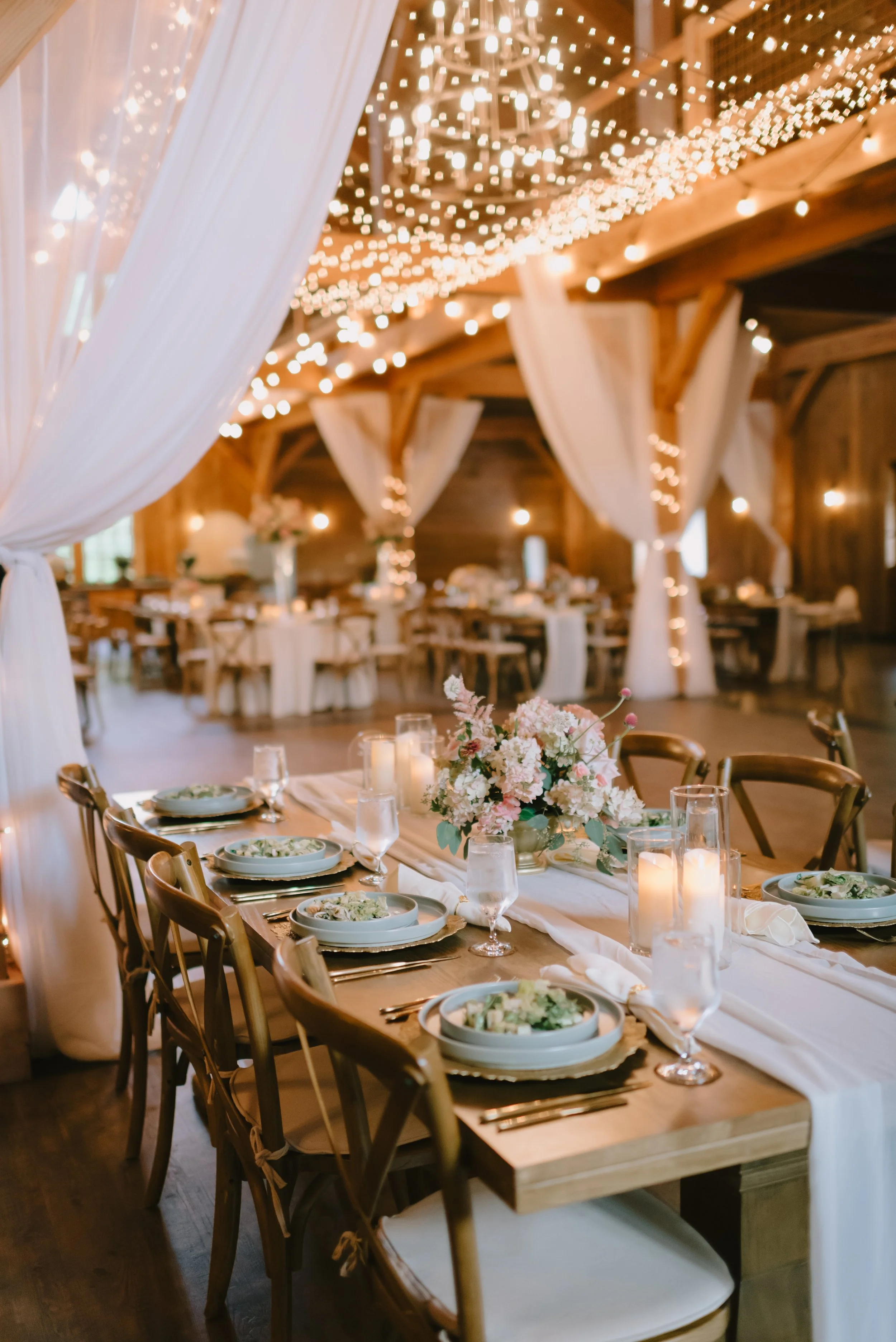 A decorated dining area with a long table set with plates, glasses, and a floral centerpiece, under string lights and draped curtains, in a rustic wooden venue.