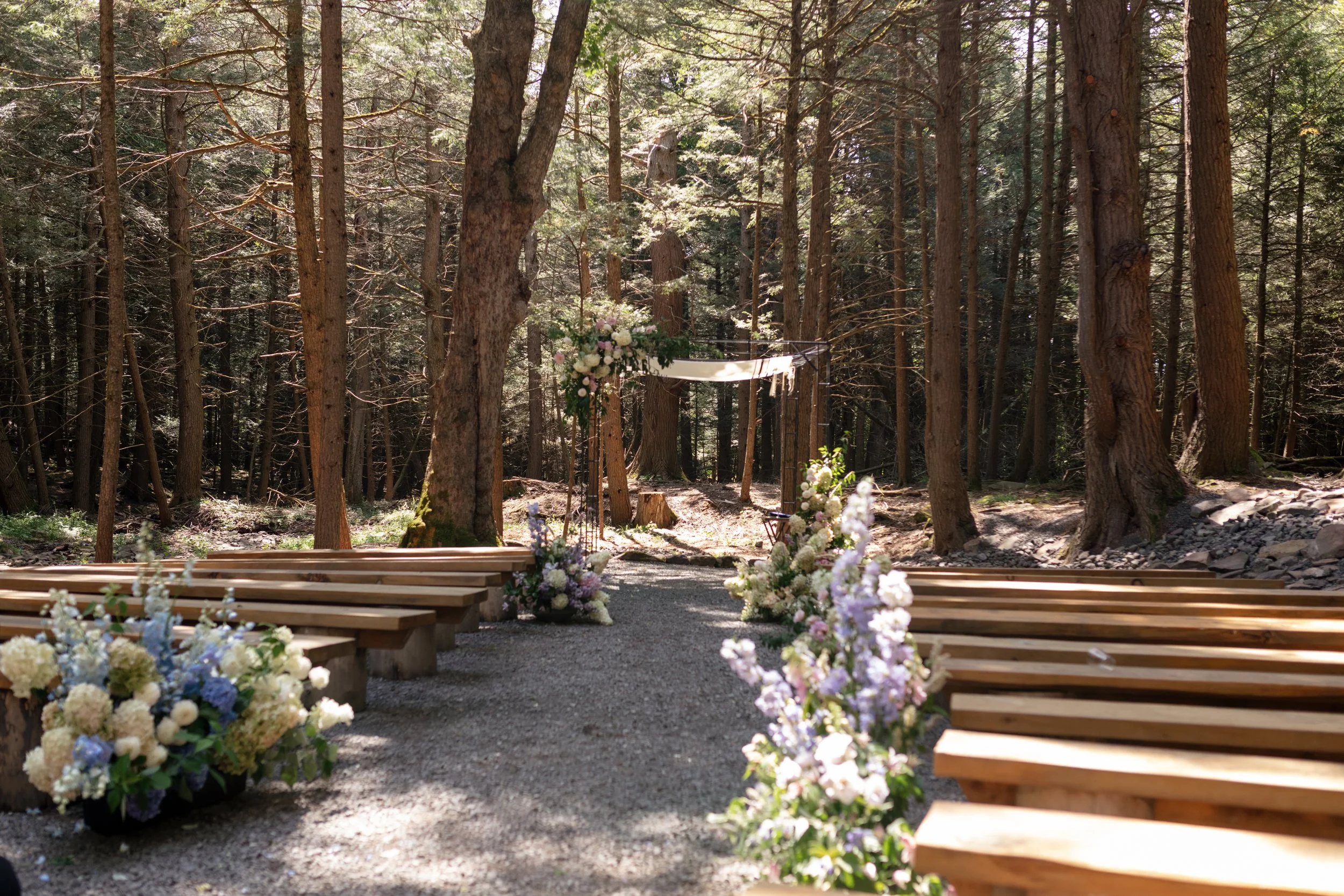 A wedding ceremony setup in a forest with wooden benches and floral arrangements along the aisle.