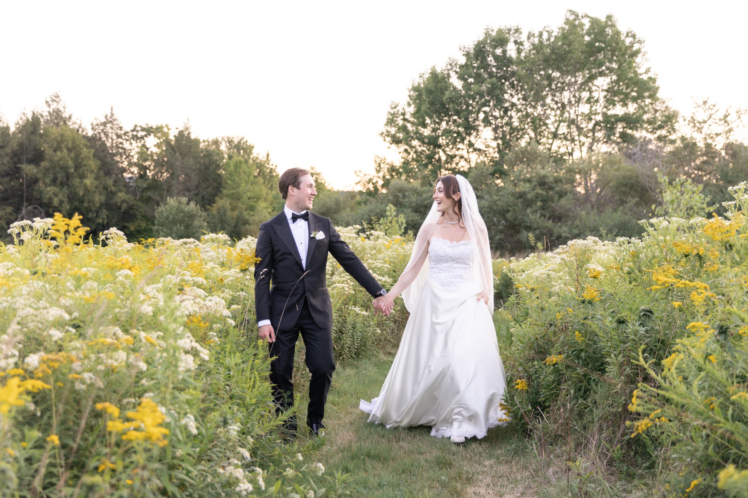 A bride and groom holding hands and smiling while walking in a yellow flower field.
