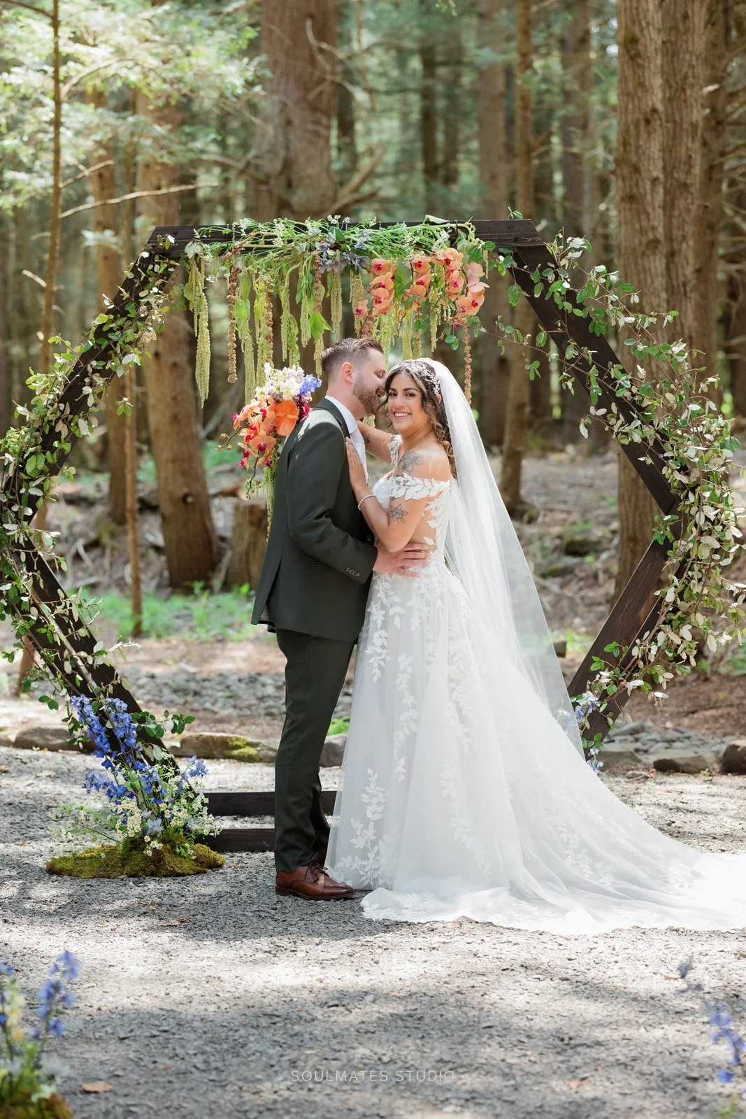 A bride and groom standing together under a floral wedding arch in a forest, smiling and embracing.