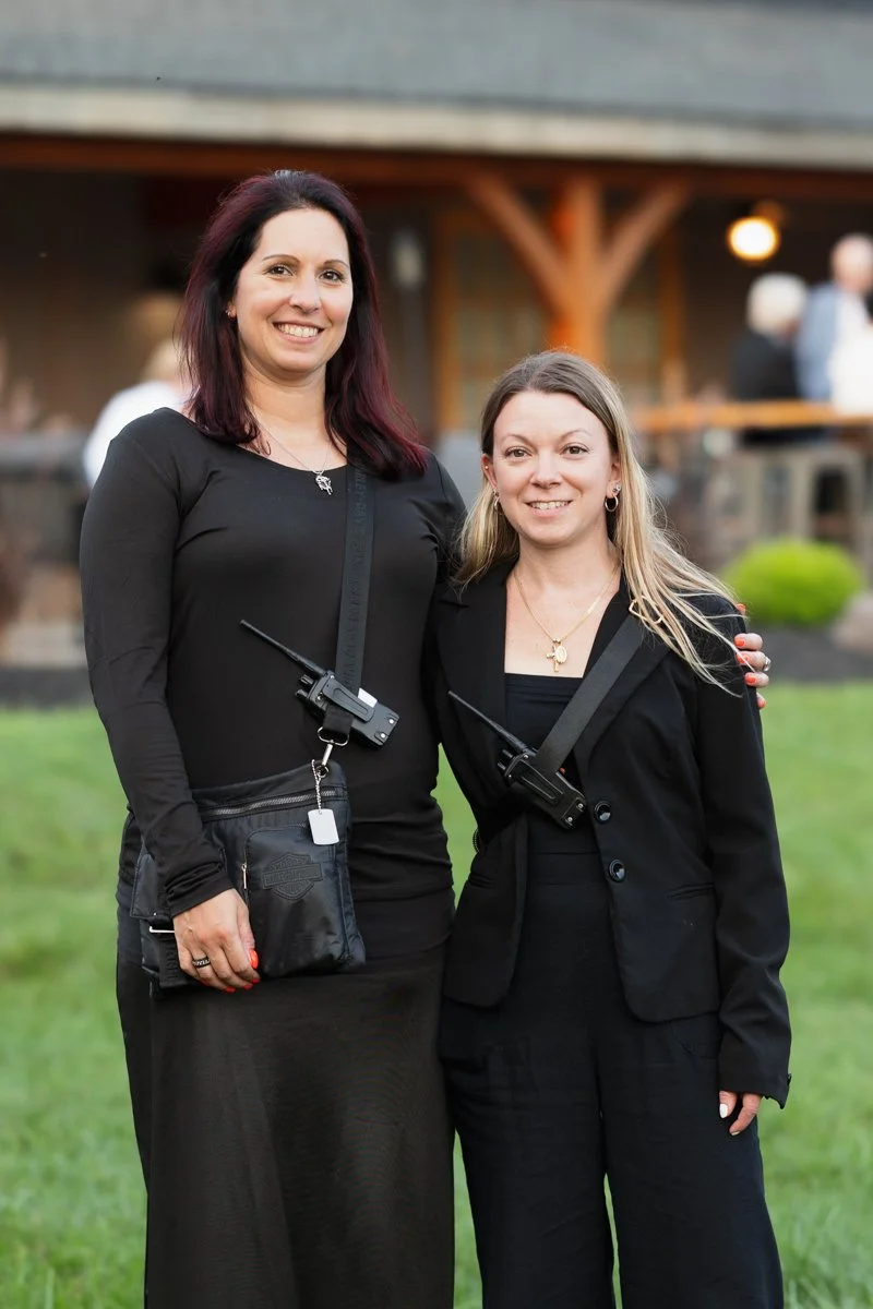 Two women standing outdoors in front of a building, wearing black outfits with radio communication devices attached to their clothing, smiling at the camera.