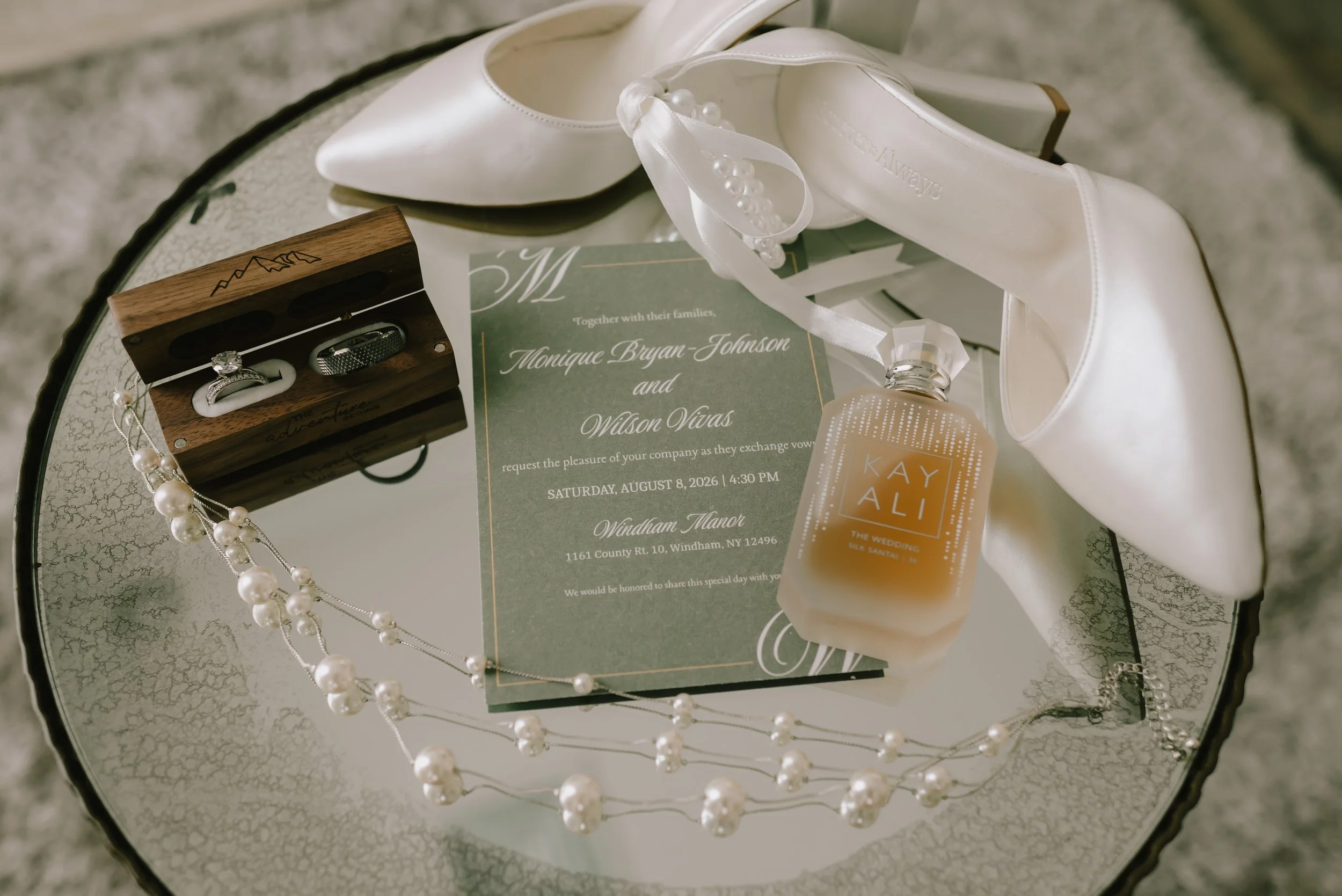 White satin wedding shoes with a pearl and ribbon decoration, placed on a glass tray. A pearl necklace, a wedding invitation with green and white design, a bottle of Kayali perfume, and a wooden box with jewelry inside are also arranged on the tray.