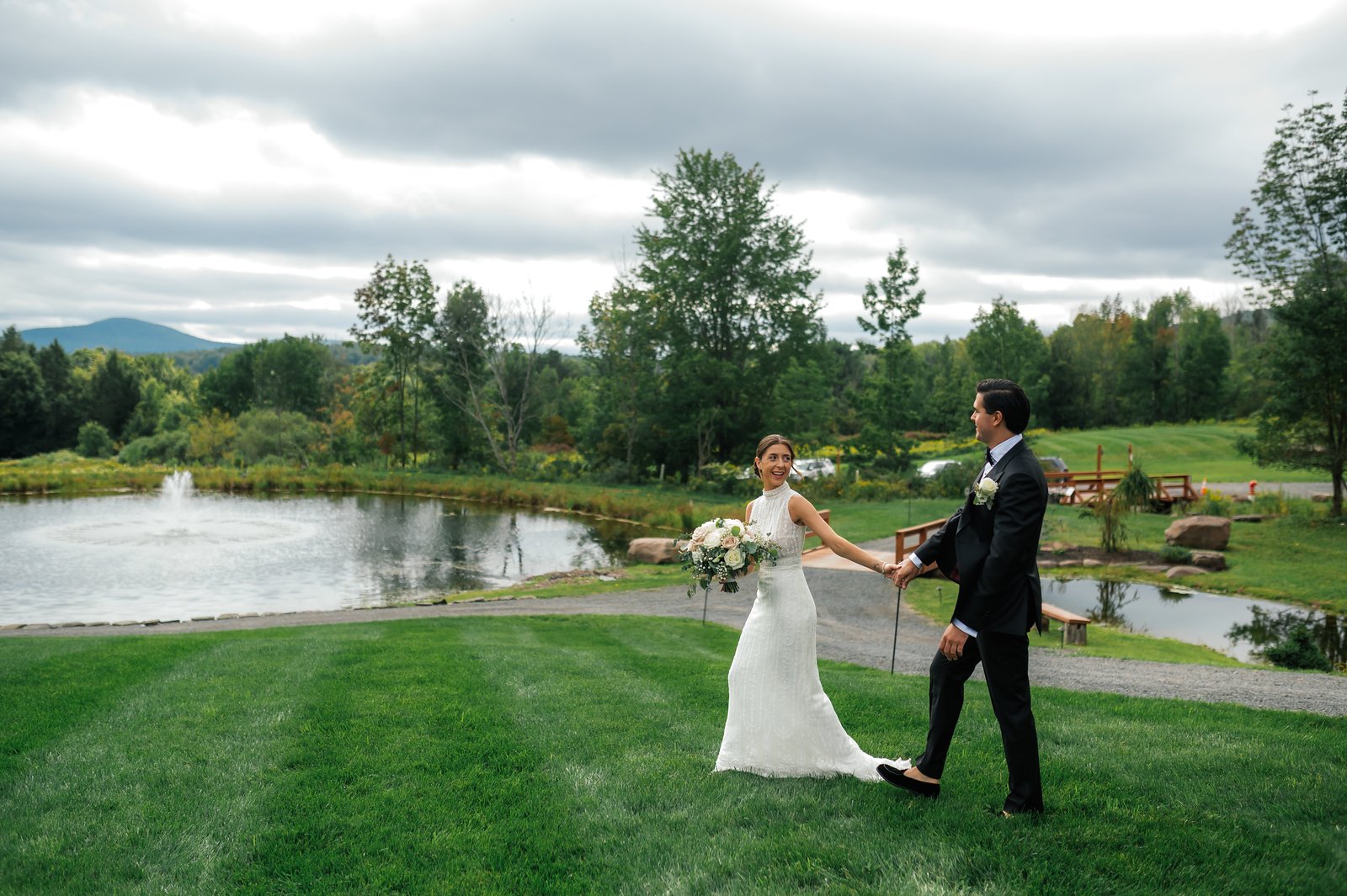 A bride and groom holding hands and walking on the grass near a pond during a wedding photoshoot. The bride is in a white wedding dress holding a bouquet, and the groom is dressed in a black suit. There are trees and a cloudy sky in the background.