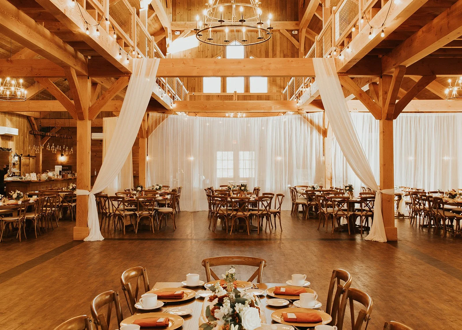 Indoor event space with wooden beams and walls, decorated for a celebration with tables set with orange napkins, white plates, teacups, floral centerpieces, and draped white fabric. There are overhead chandeliers and string lights adding a warm ambiance.