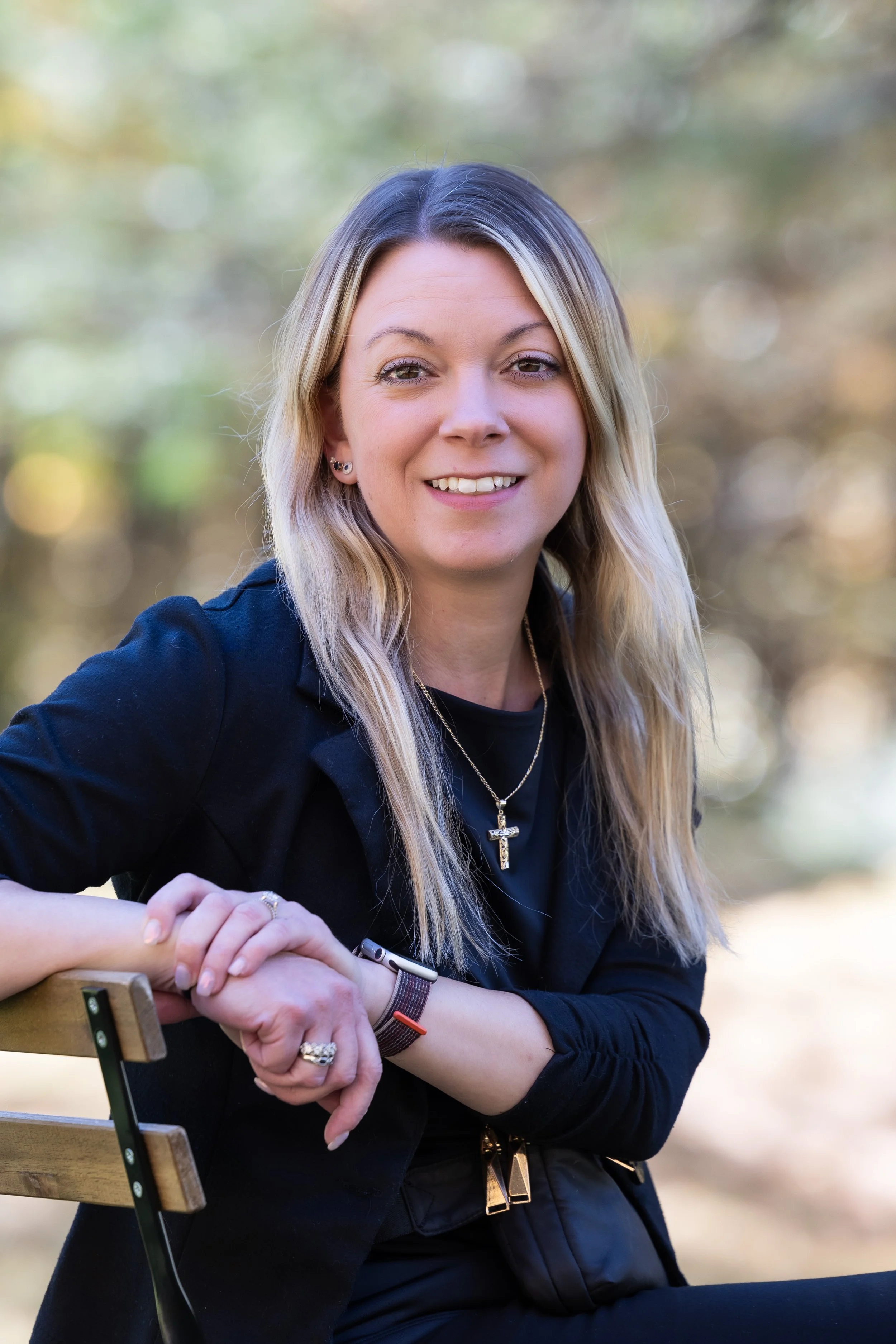 A woman with long blonde hair, wearing a black blazer and jewelry, sitting on a park bench with blurred trees in the background.