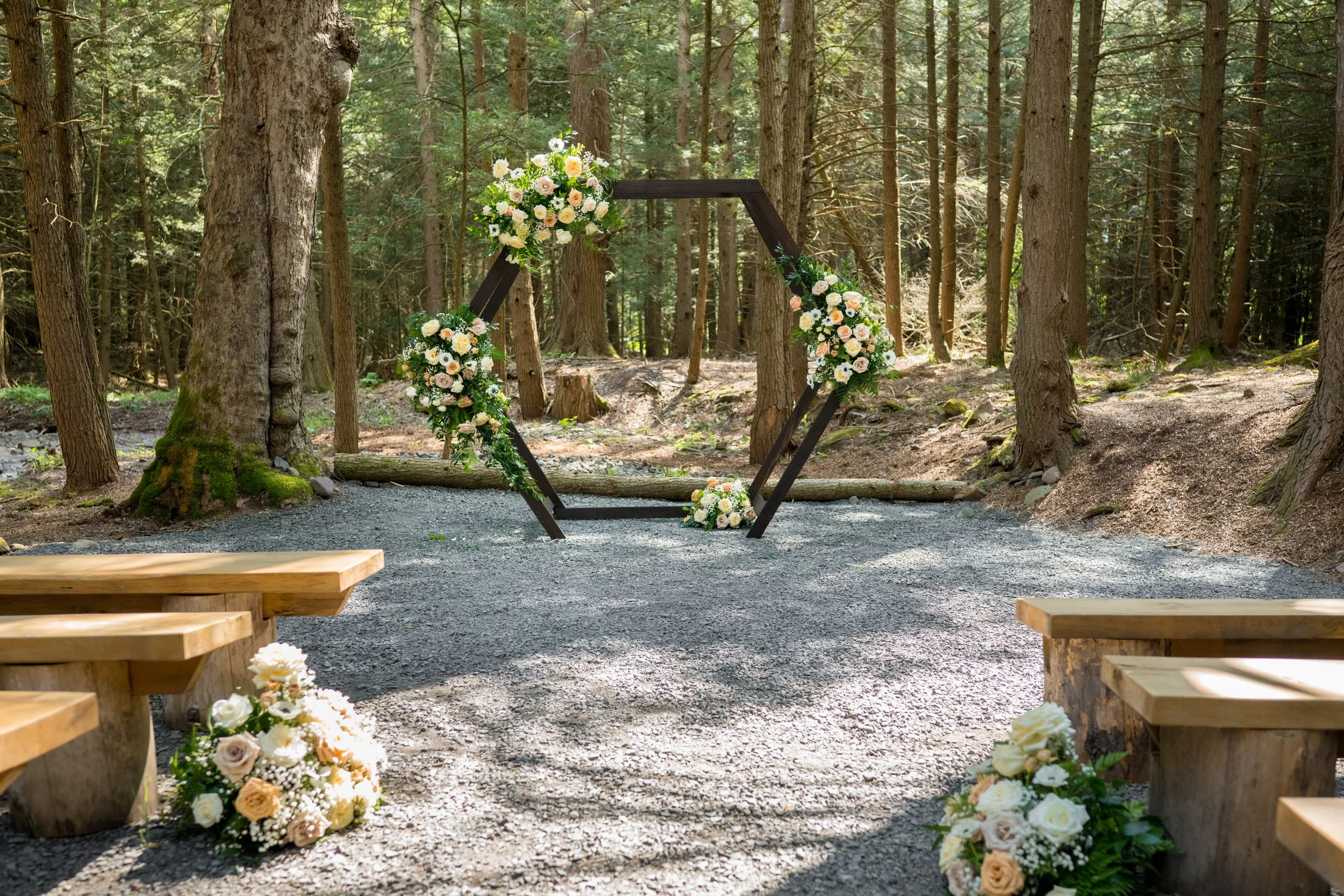 A wedding arch decorated with white and peach flowers is set up outdoors among tall trees in a forest.