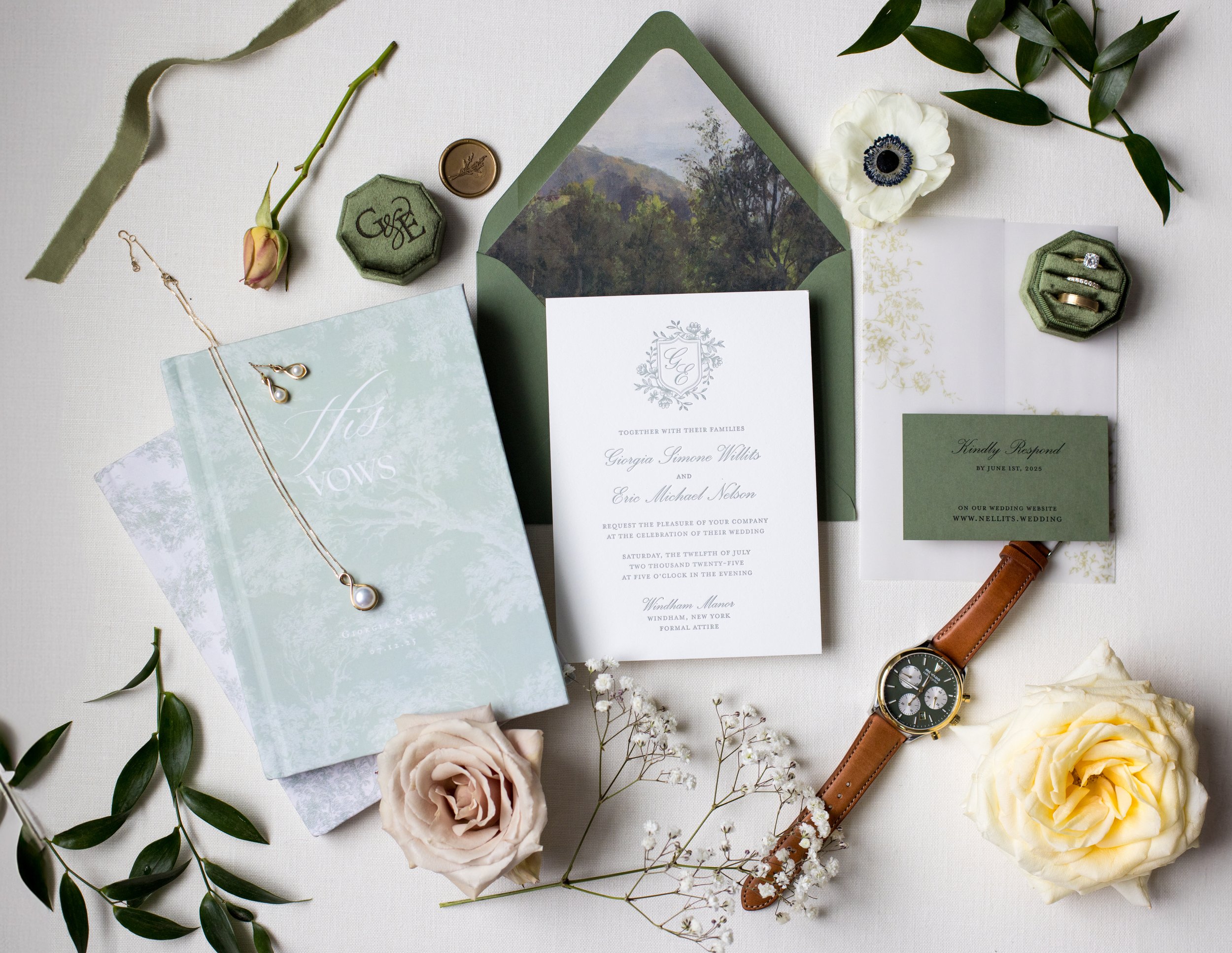 Wedding invitation suite with greenery, flowers, jewelry, watch, and rings on white table.