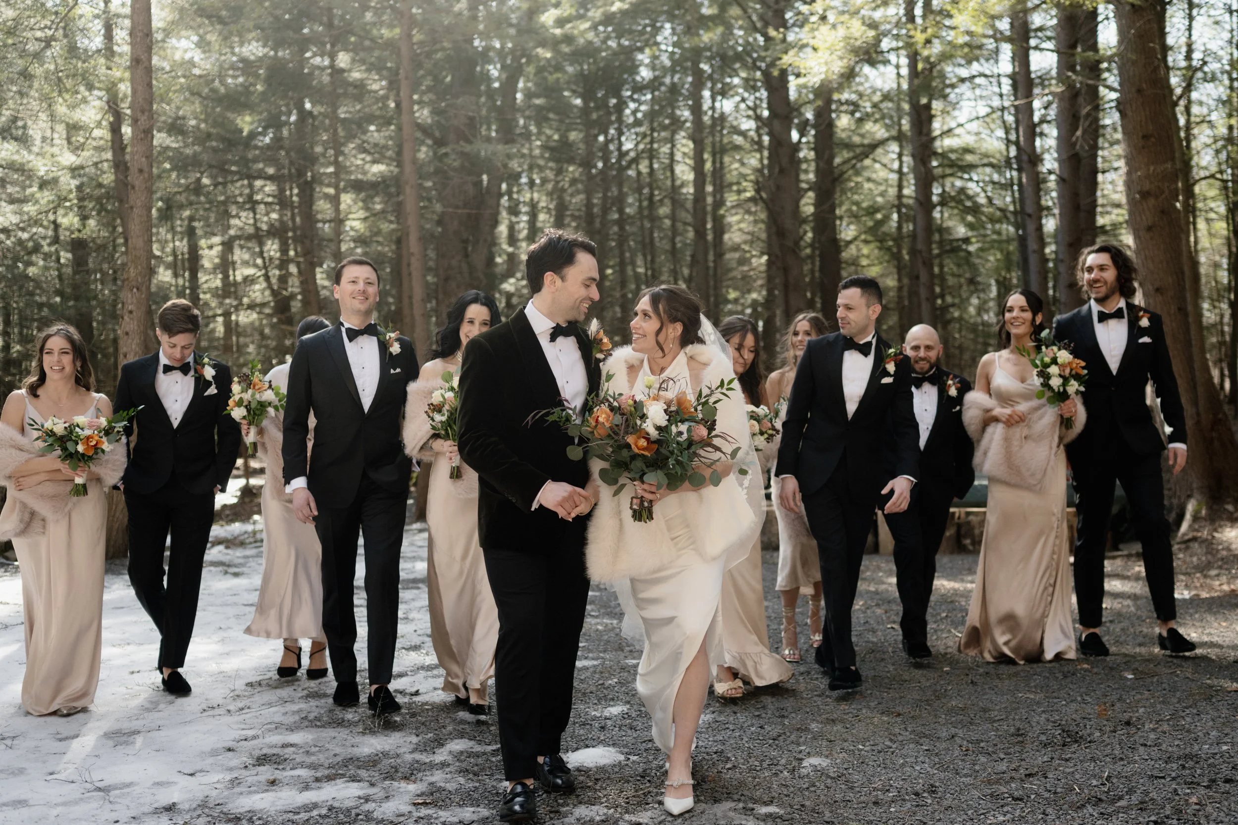 A group of wedding guests, including a bride and groom, walk through a snowy forested area with tall trees, dressed in formal attire and holding bouquets of flowers.