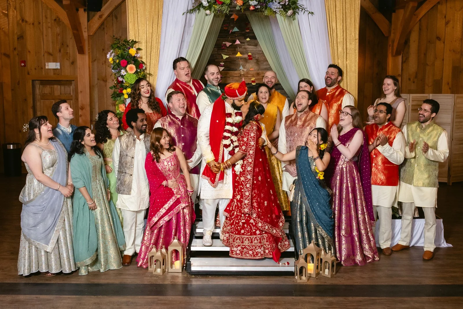 A group of people in traditional Indian wedding attire celebrating a wedding ceremony, with the bride and groom in the center, in a decorated indoor venue.
