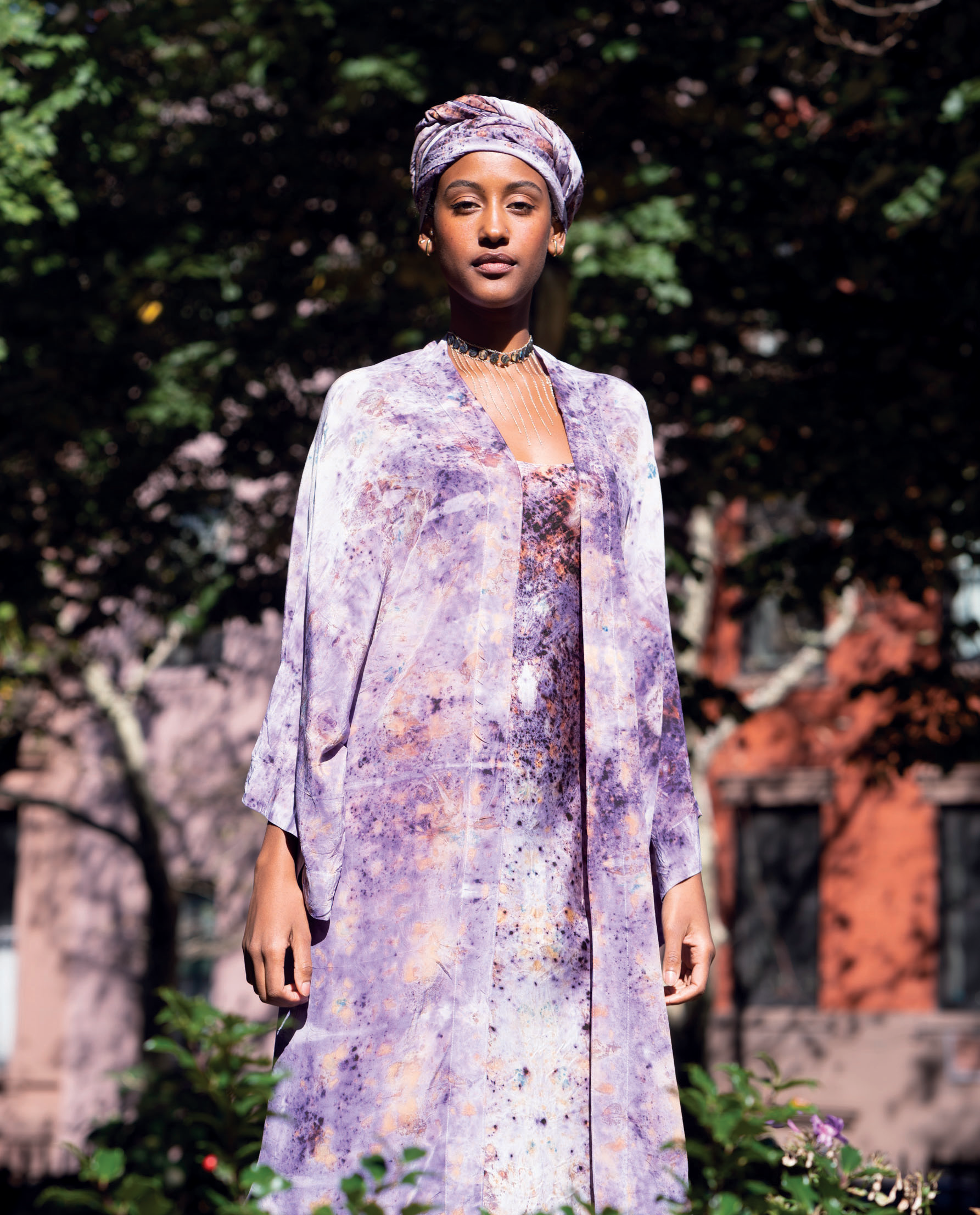 A woman dressed in a lavender and purple patterned outfit with a matching headwrap, standing outdoors with trees and a brick building in the background.