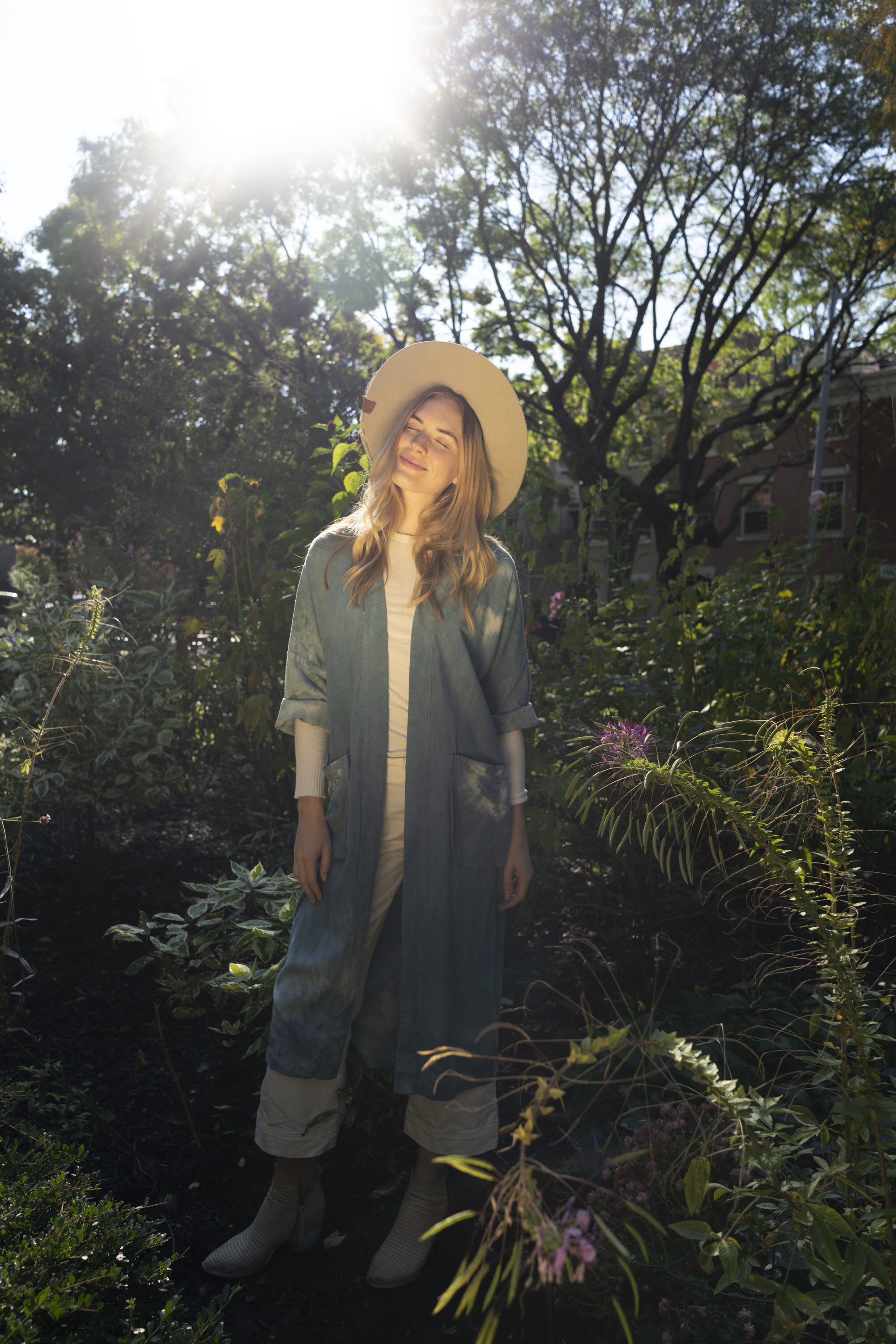 A woman with long blonde hair smiling with her eyes closed, wearing a wide-brimmed hat and a blue coat, standing outdoors in a garden with sunlight streaming through the trees.
