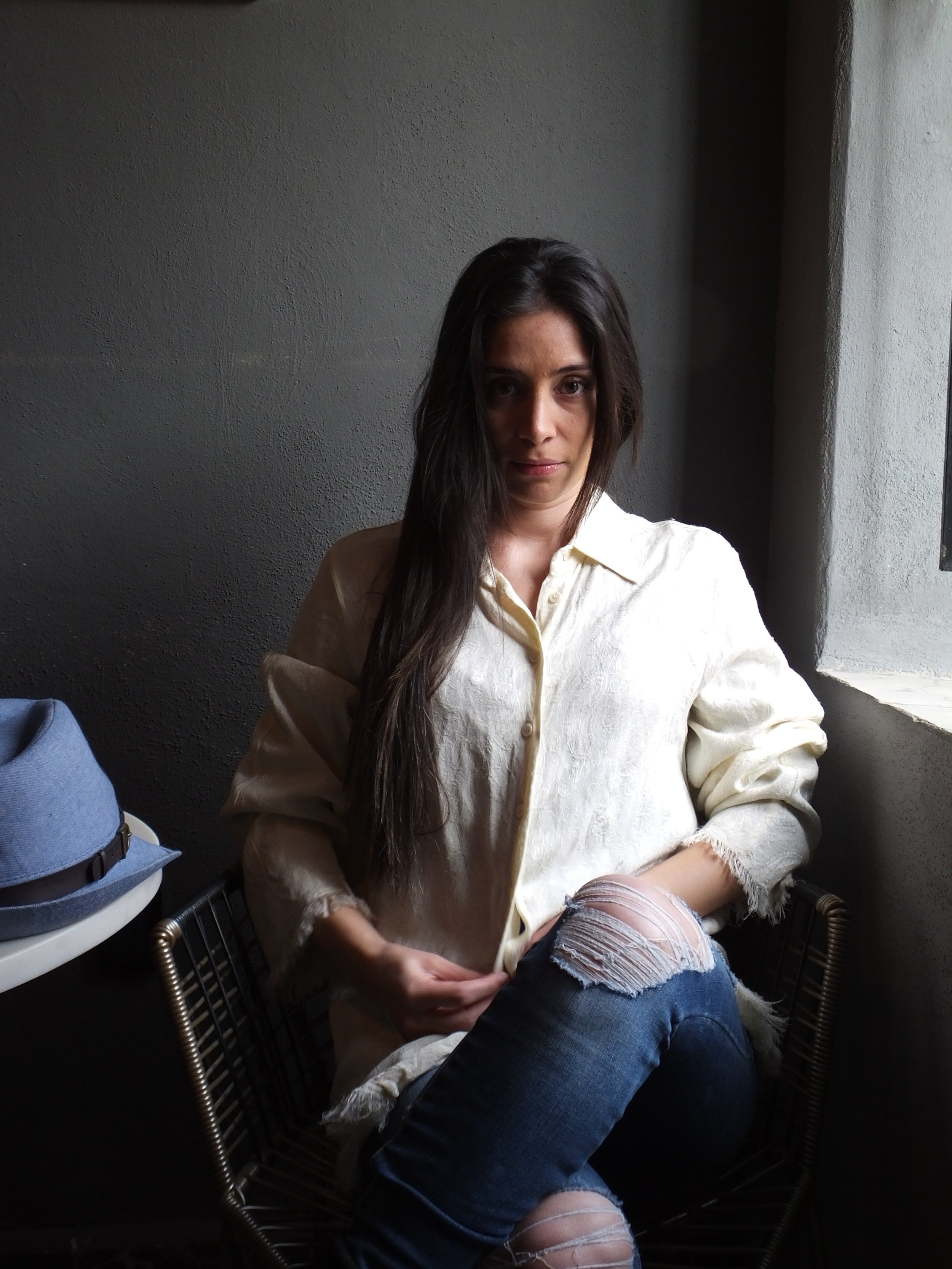 A woman with long dark hair wearing a cream-colored oversized shirt and ripped jeans, sitting on a wire chair near a window with natural light.