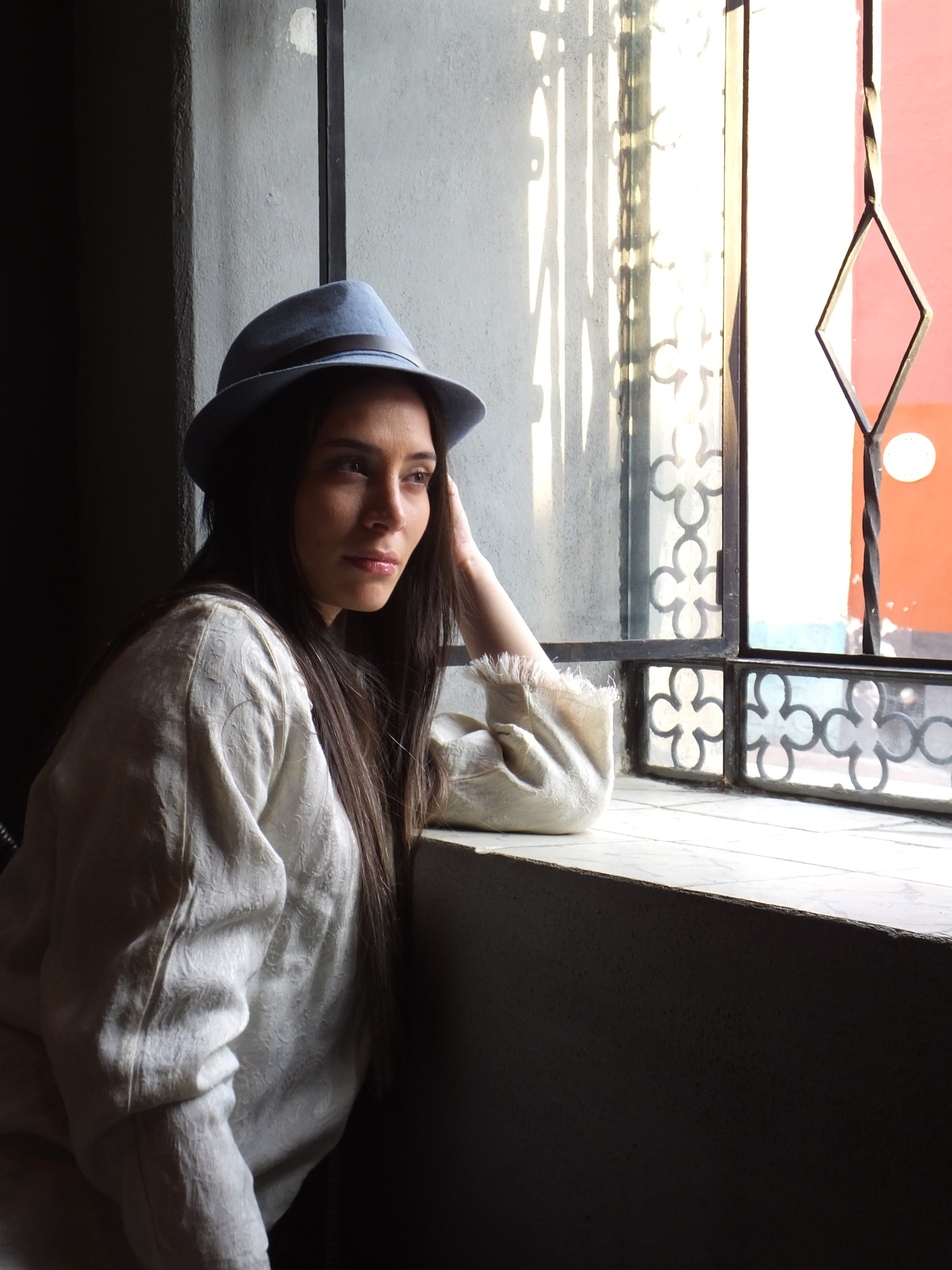 A young woman with long dark hair, wearing a gray hat and a white textured sweater, sitting by a window with decorative ironwork, looking outside thoughtfully.