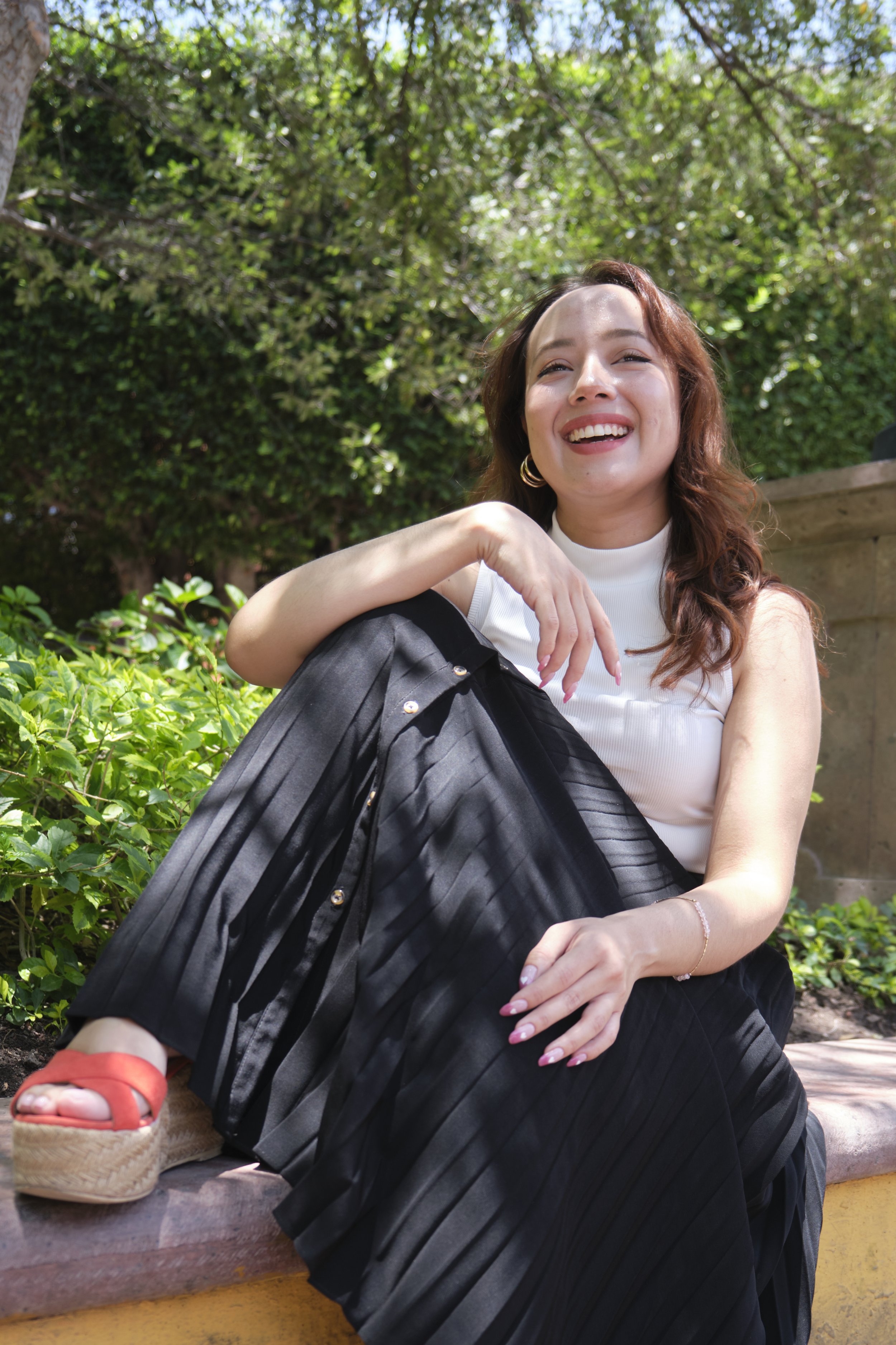 A woman with wavy brown hair, wearing a white sleeveless top, black pleated skirt, and red wedge sandals, sitting on a bench outdoors surrounded by lush green foliage while smiling joyfully.