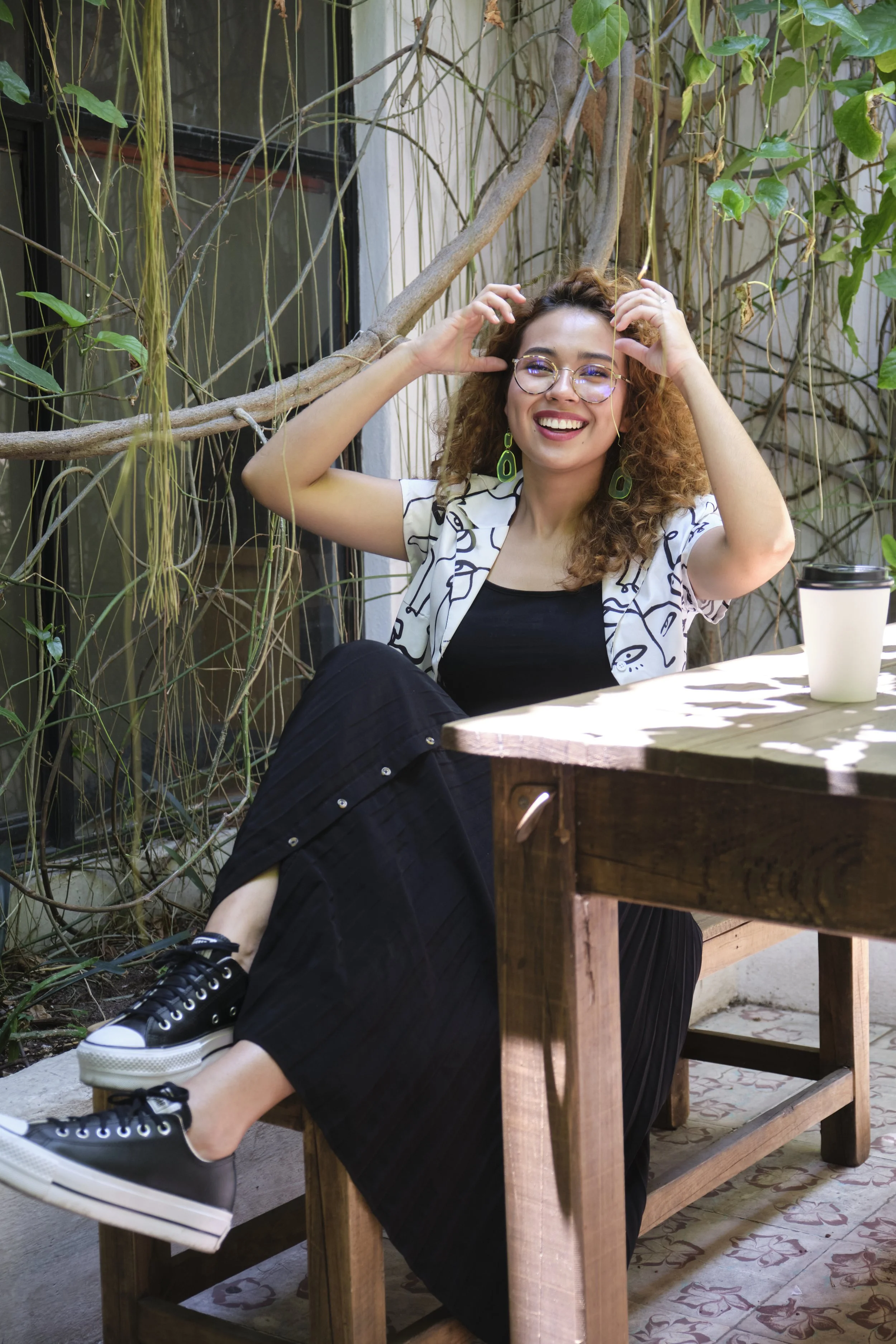 A woman with curly hair, glasses, and green earrings smiling and sitting outdoors at a wooden table surrounded by vines and plants.