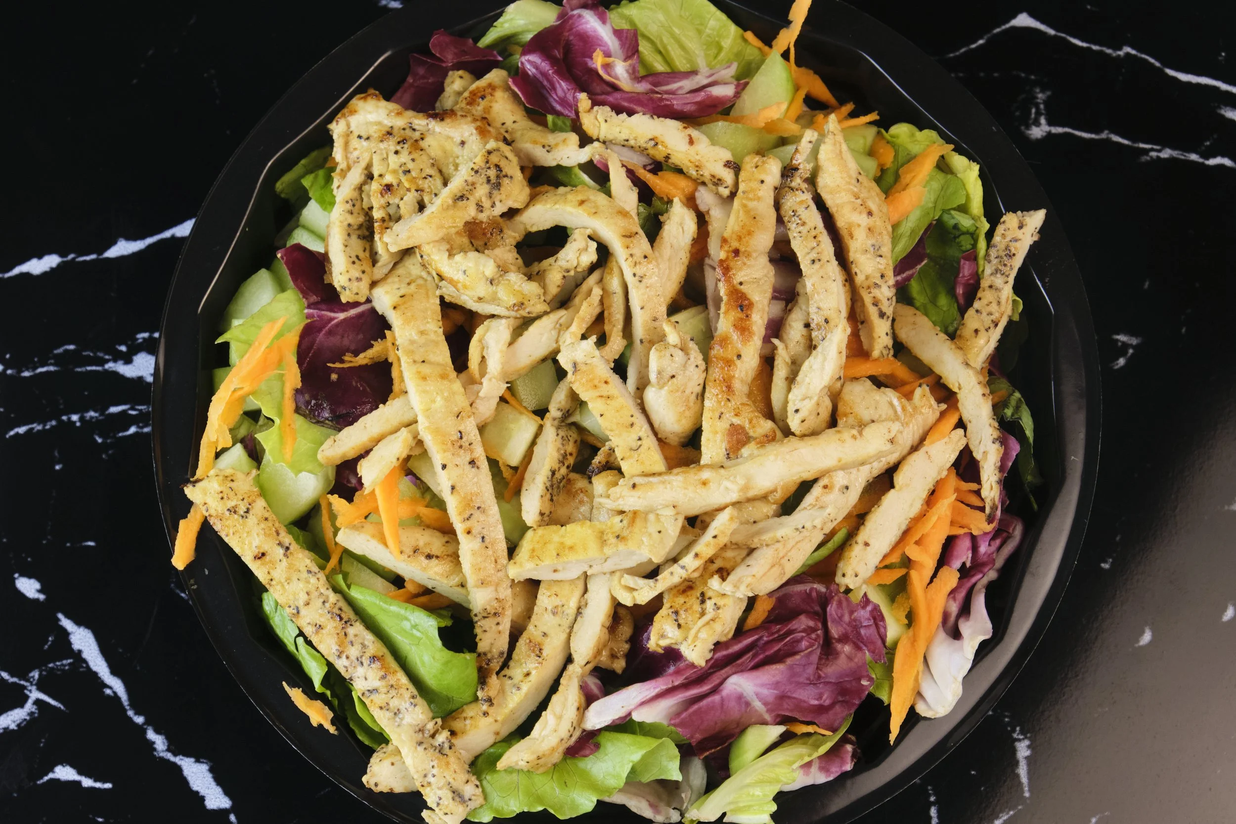 Salad with grilled chicken strips, shredded carrots, cucumbers, and mixed greens in a black bowl on a black and white marble surface.