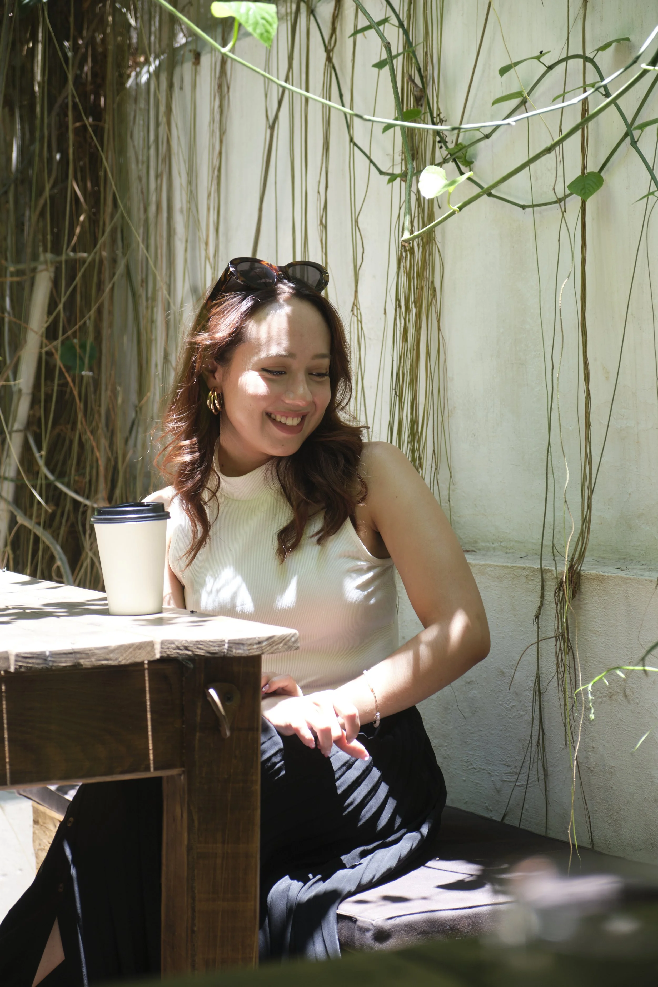 A woman with brown hair, sunglasses on her head, sitting outdoors at a wooden table with a coffee cup, smiling, surrounded by hanging plant vines on a bright sunny day.