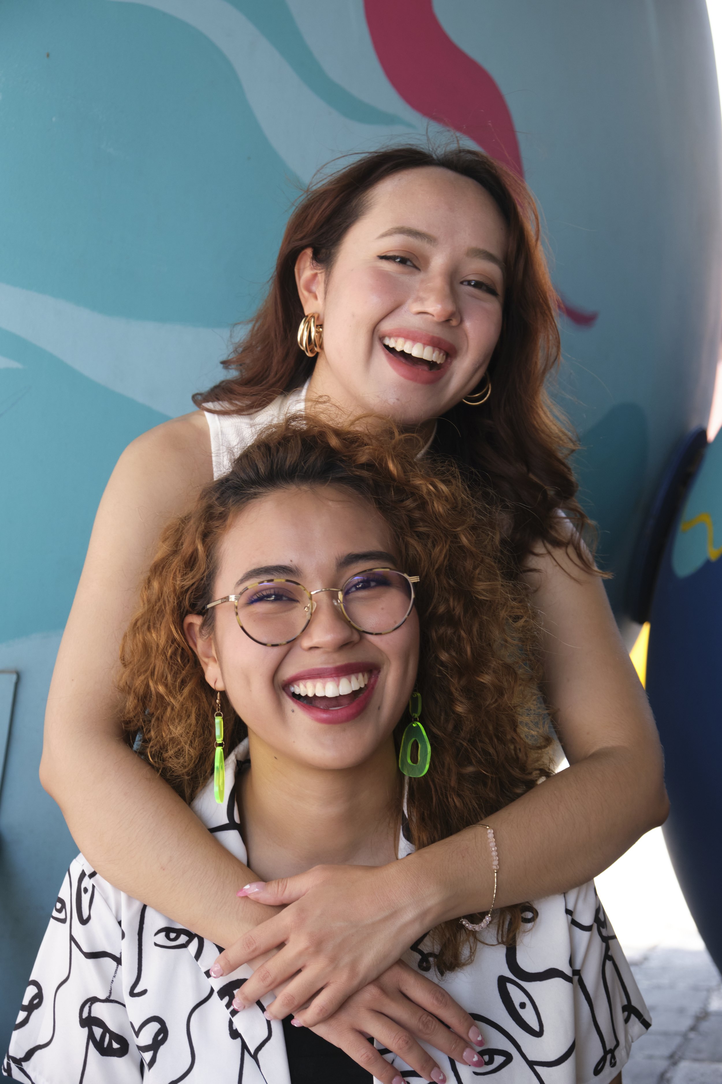 Two sisters smiling and hugging each other, one standing behind the other, in front of a colorful mural.