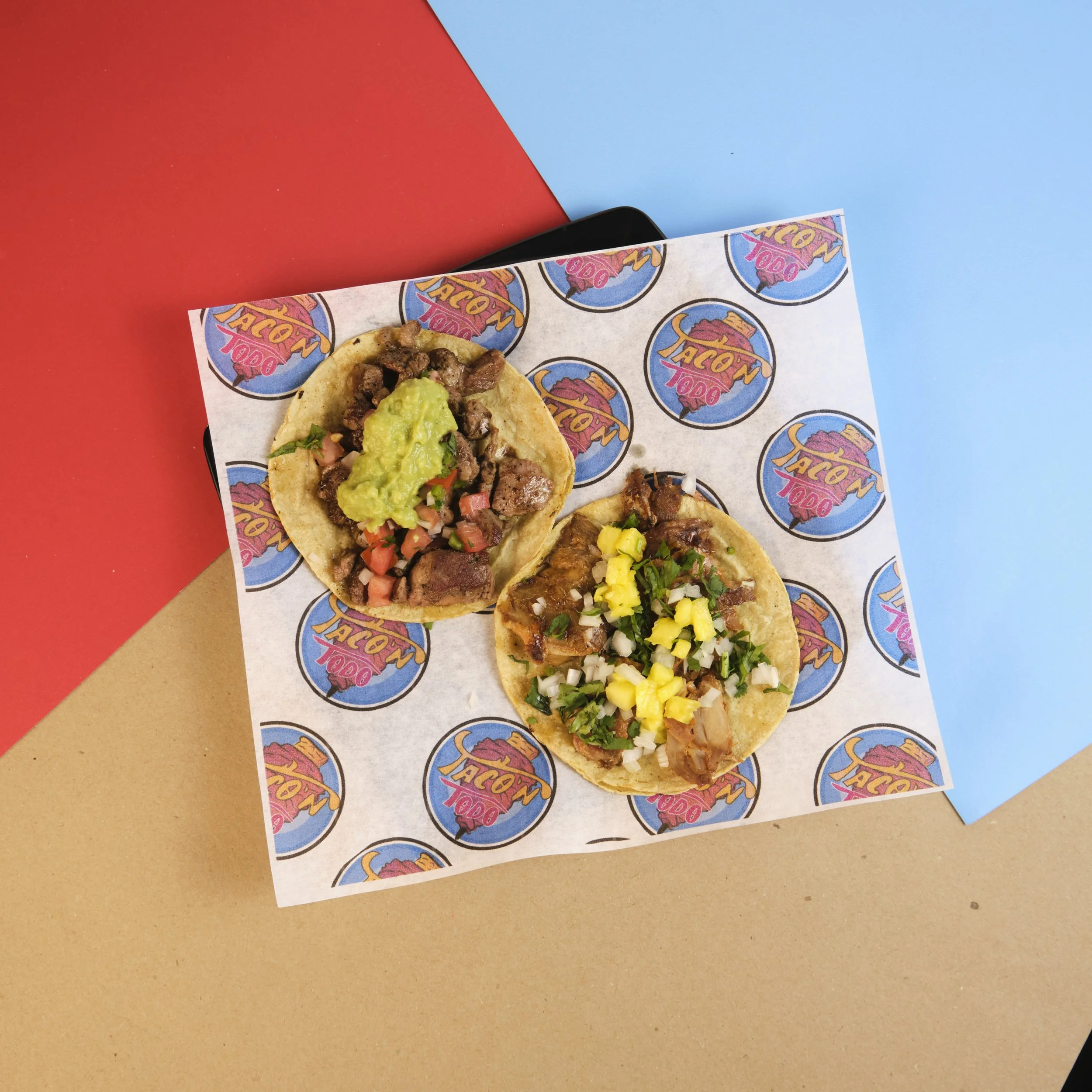 Two tacos with various toppings on a taco-shaped paper, placed on a multicolored background.