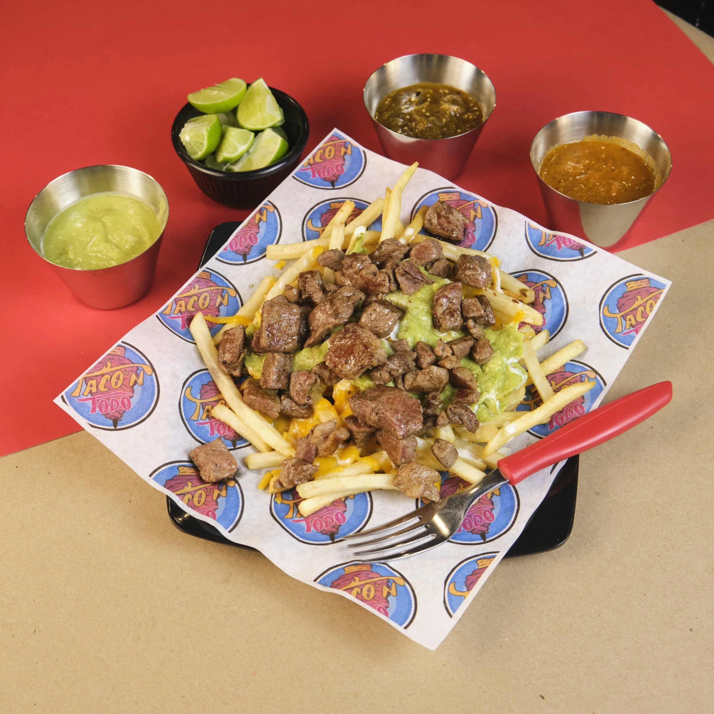 A plate of nachos topped with beef, melted cheese, and guacamole, served on branded paper; accompanied by lime wedges, two cups of salsa, and green guacamole sauce, on a red and tan table.