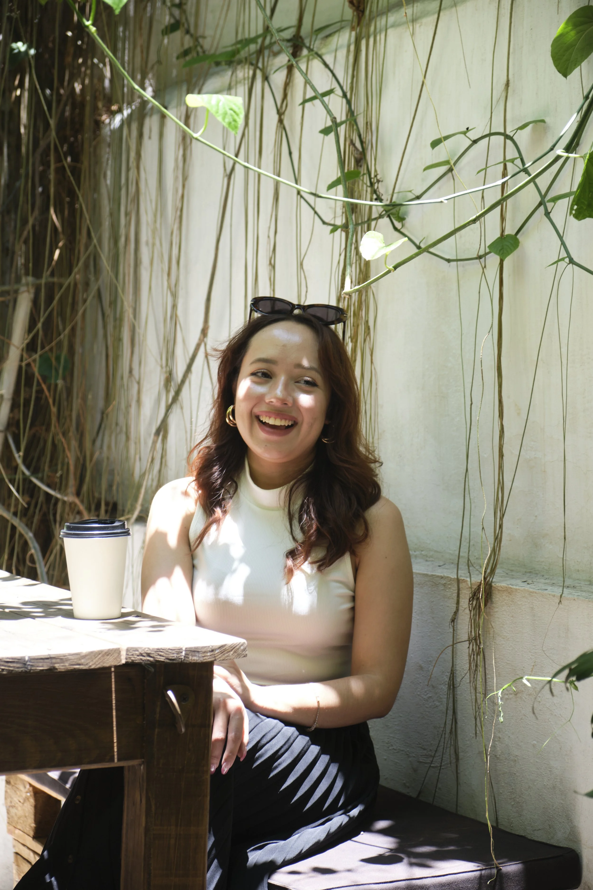 A woman with wavy brown hair and sunglasses on her head, smiling while sitting outdoors at a wooden table with a disposable coffee cup, surrounded by green plants and vines.