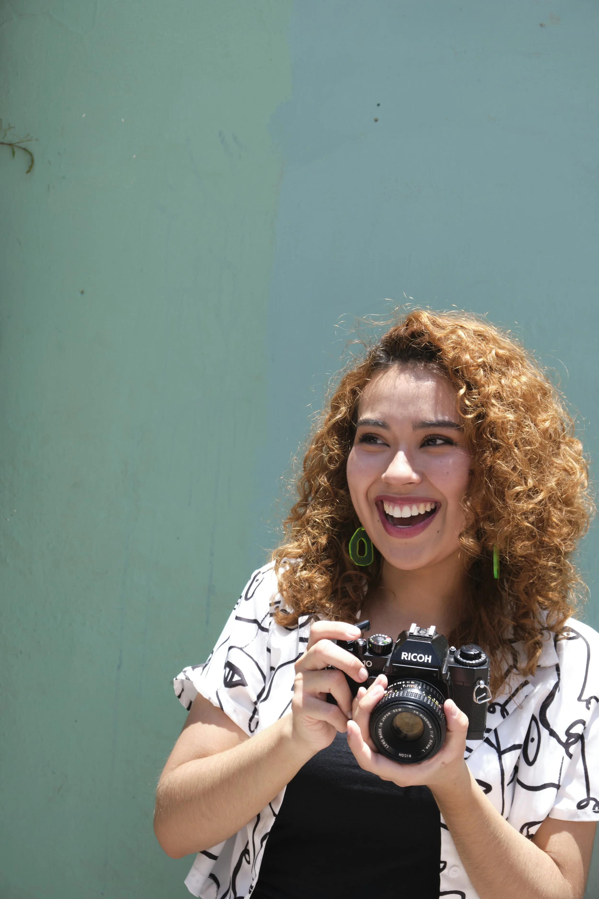 A woman with curly red hair wearing a black and white patterned shirt, smiling and holding a Ricoh camera, standing in front of a light green wall.
