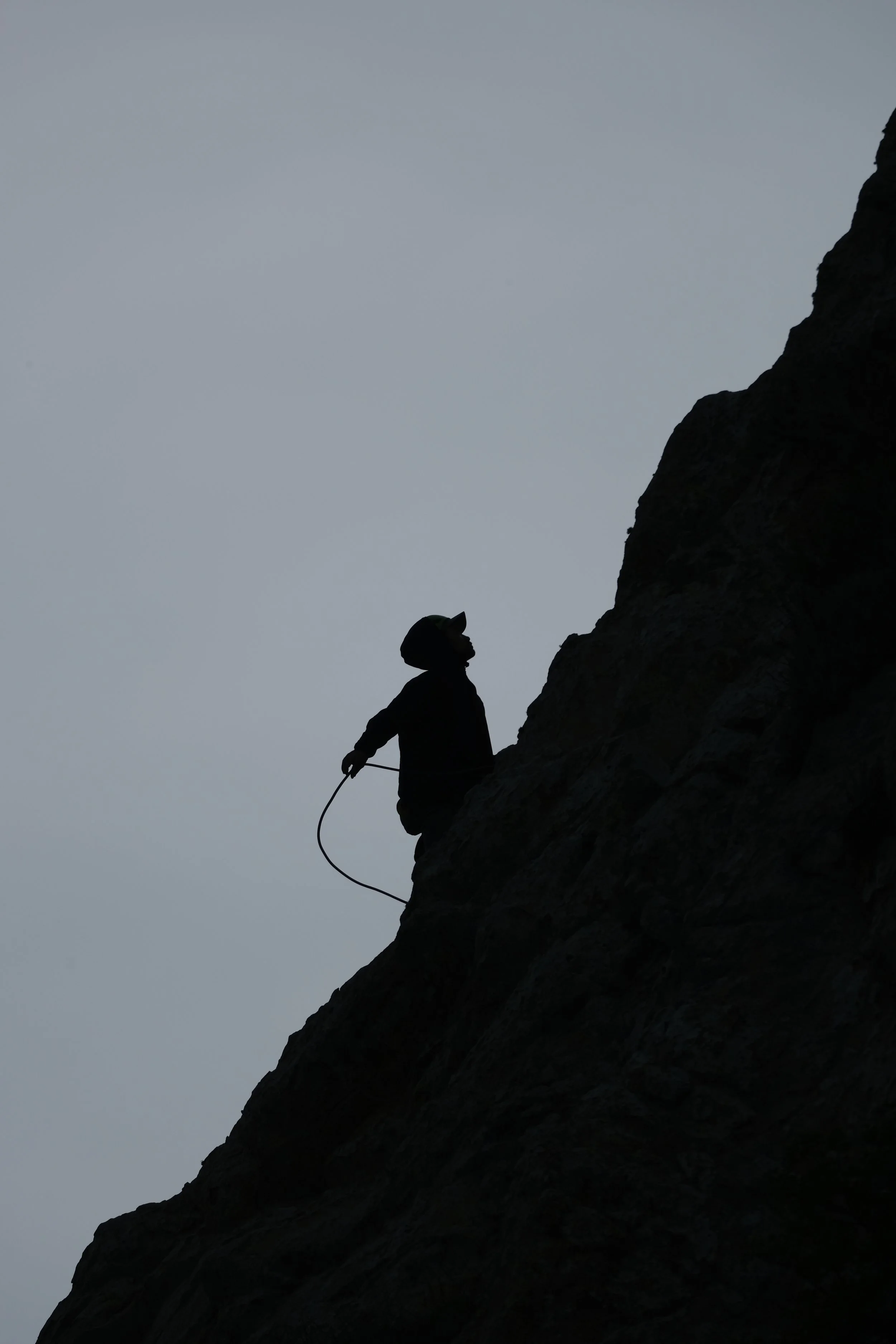 Silhouette of a person climbing a steep rock face, holding a safety rope, against a cloudy sky.